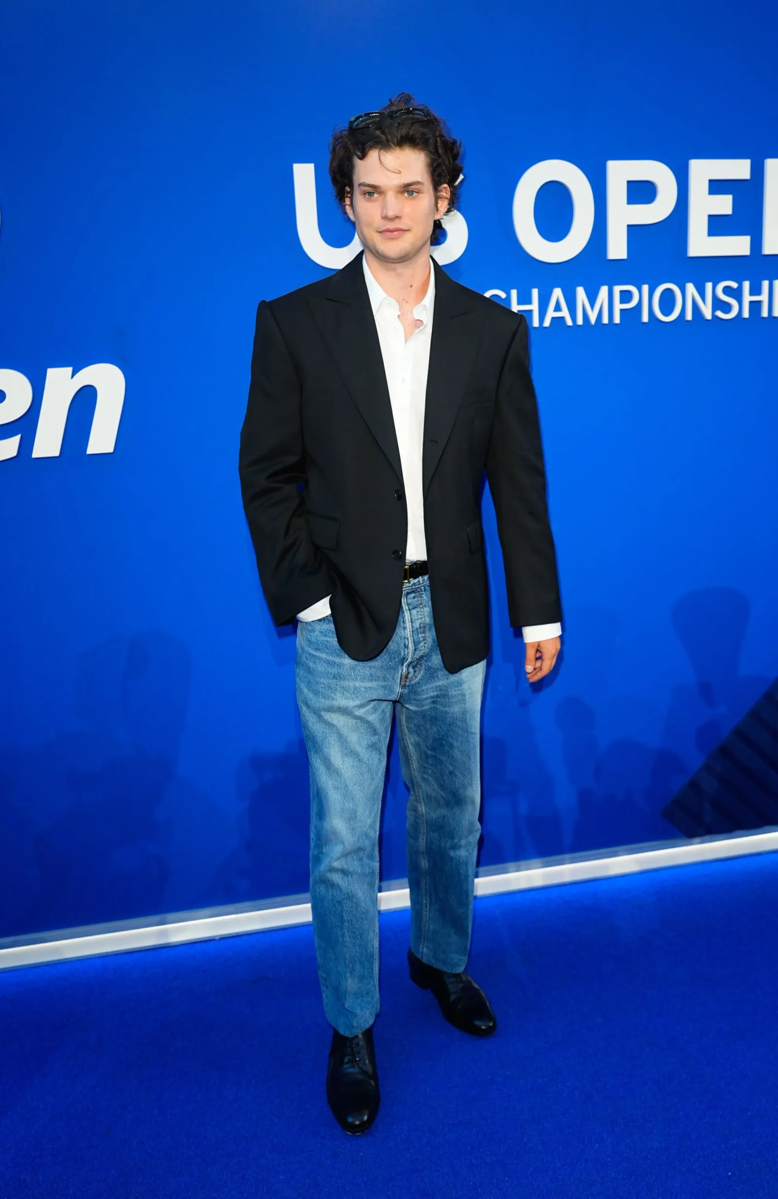 Young man in a black blazer and white shirt stands against a blue backdrop with 'US OPEN CHAMPIONSHIP' text nearby.