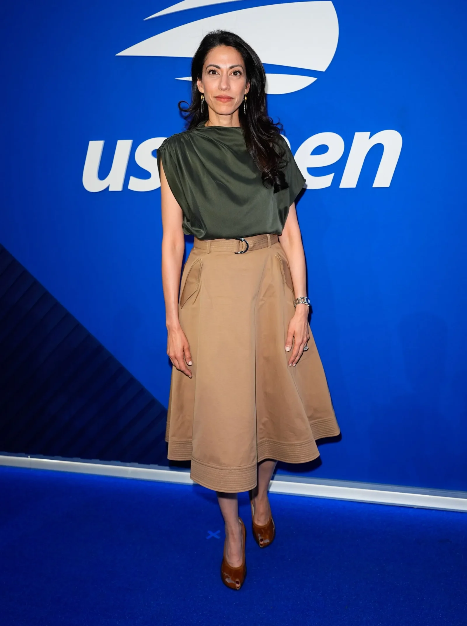 Woman in an olive green draped top and tan midi skirt posing in front of a blue backdrop with a white logo behind her.