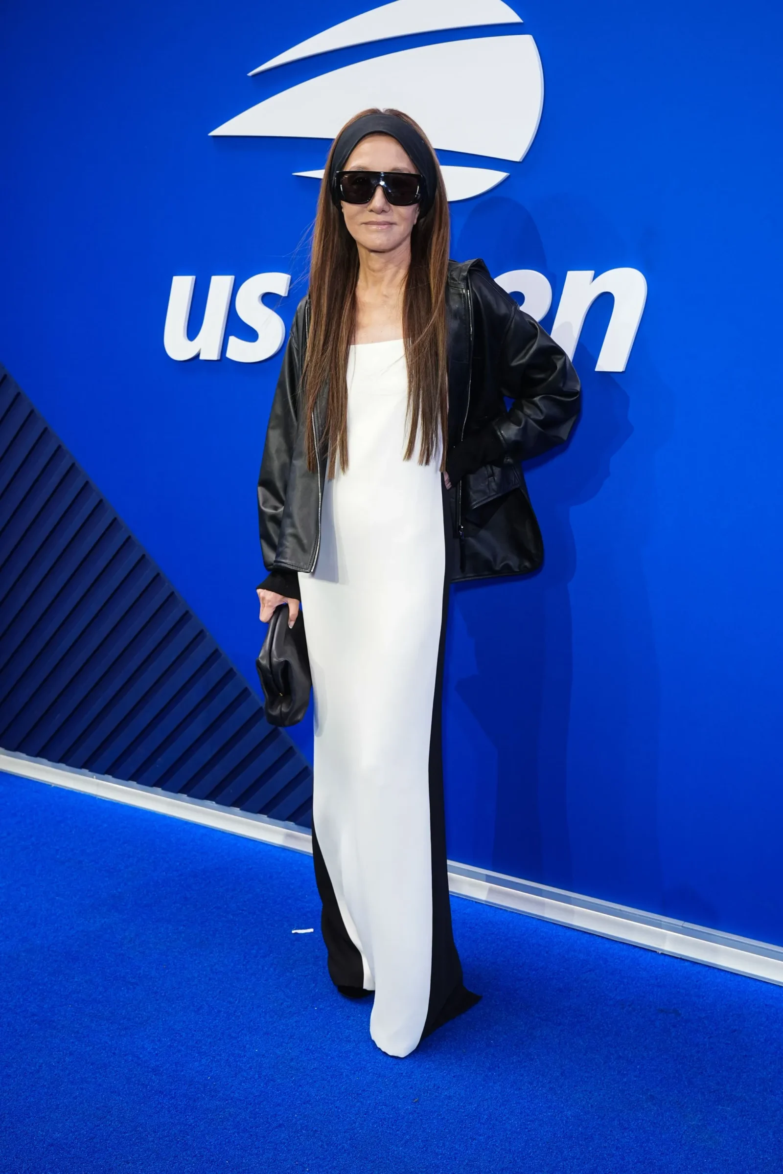 Person wearing a white dress with a black leather jacket and oversized sunglasses posing in front of a blue backdrop with a white logo partially visible (