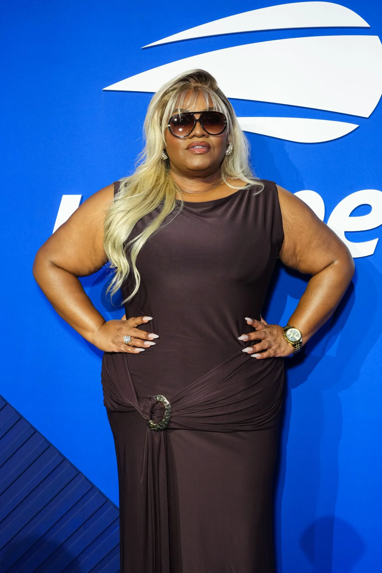 Woman with long blonde hair and sunglasses posing with hands on hips against a blue backdrop with a white logo behind her.
