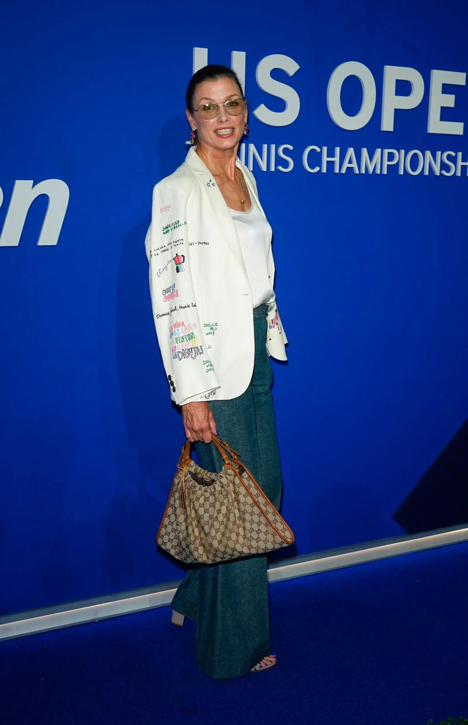 Woman in a white blazer and teal pants posing against a blue wall with 'US OPEN' text, holding a beige patterned handbag.