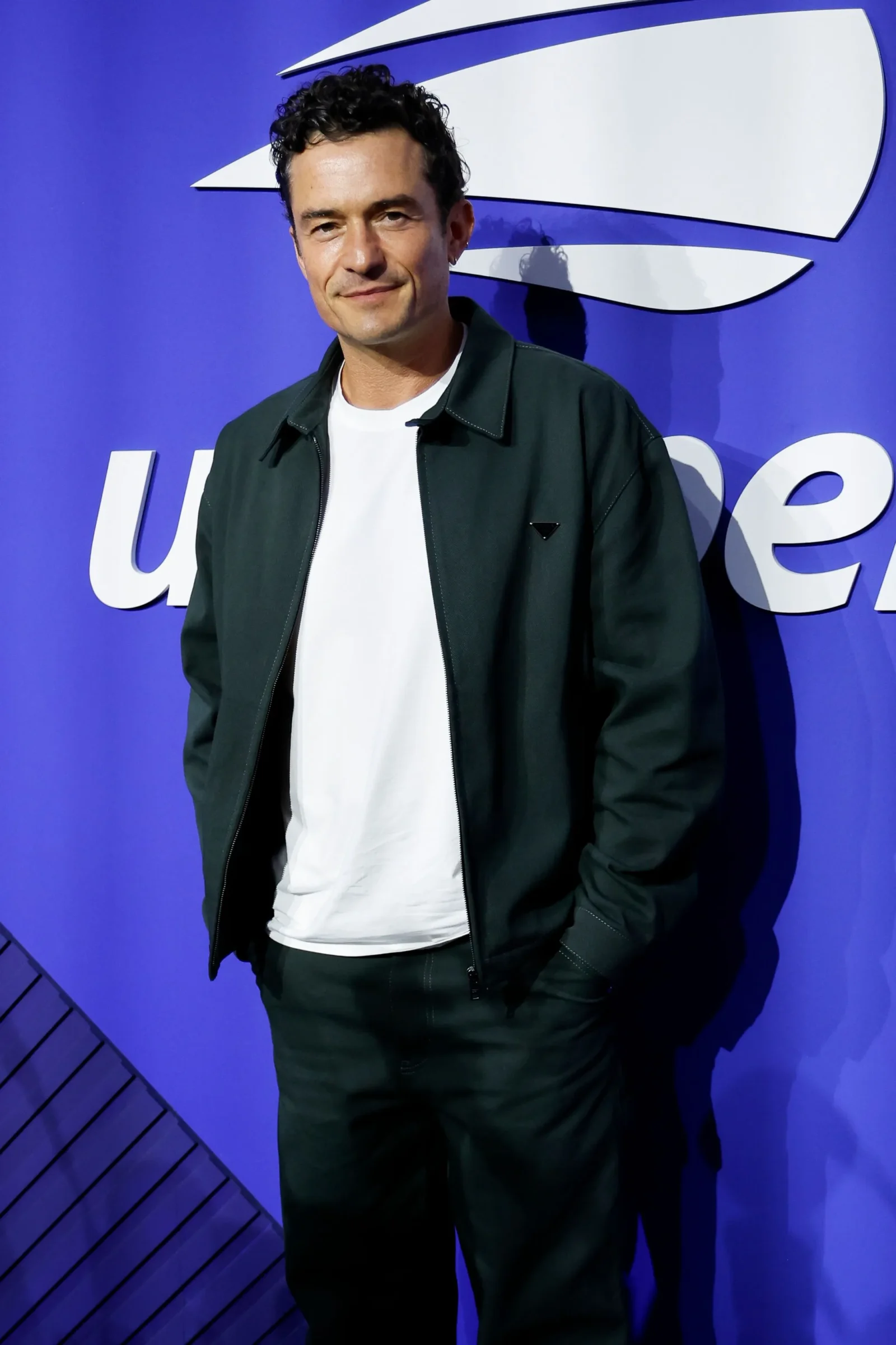 Man wearing a white t‑shirt and dark green jacket standing in front of a blue wall with a large white logo.