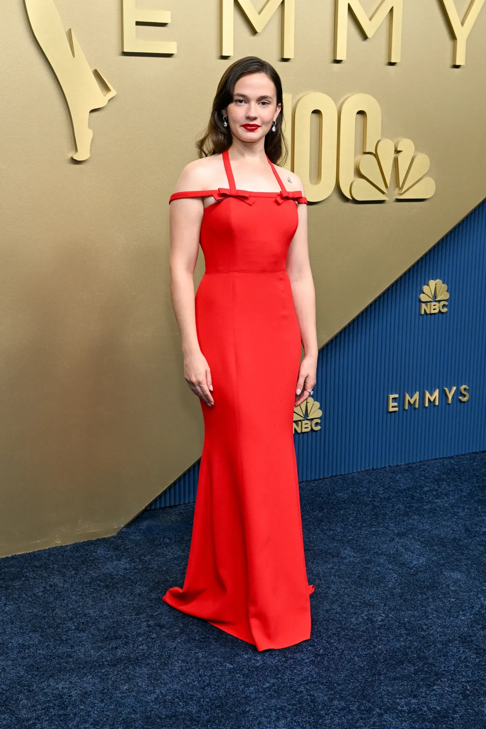 Woman in a red off-shoulder gown posing on a gold Emmy Awards backdrop with NBC logo in the background.