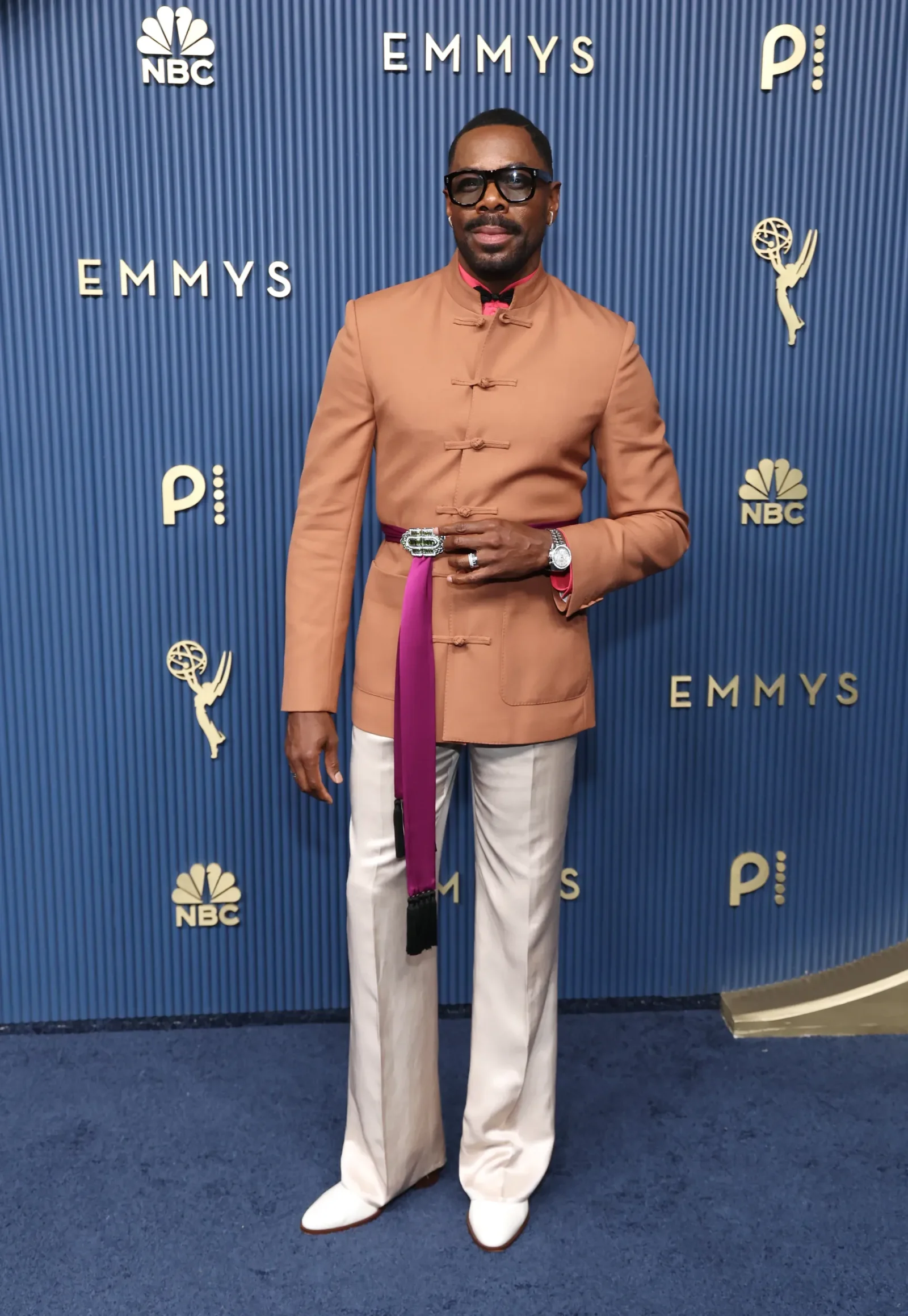 Man posing on a blue Emmy Awards backdrop in a tan mandarin-collar jacket, white pants, and a purple sash.