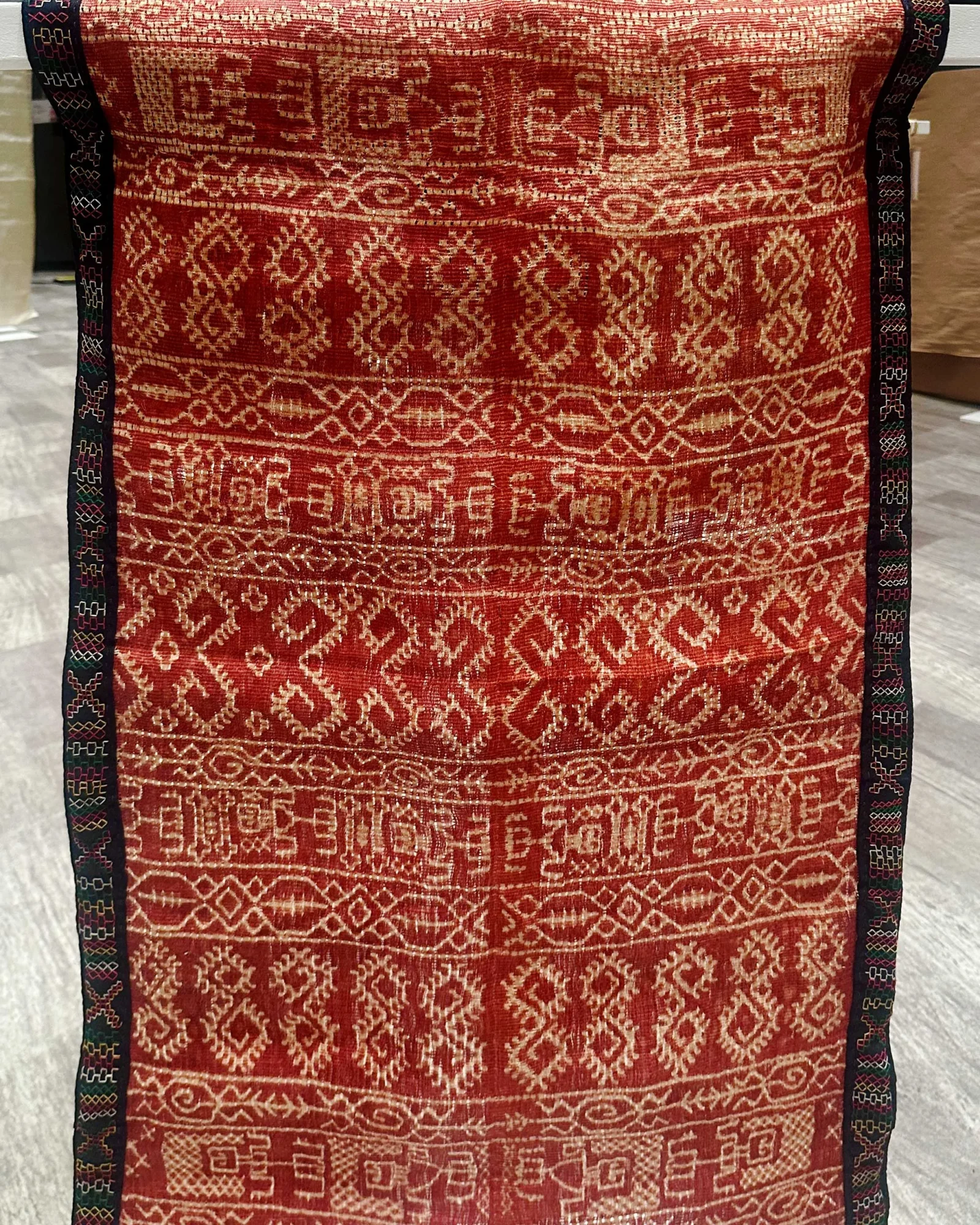 Red patterned woven rug with white geometric motifs and black decorative borders along both edges.