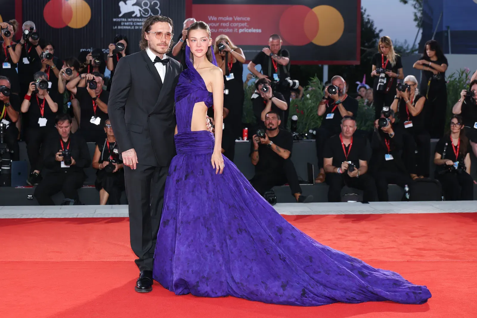 Couple posing on a red carpet; man in a black tuxedo and bow tie, woman in a dramatic purple gown with a long train surrounded by photographers