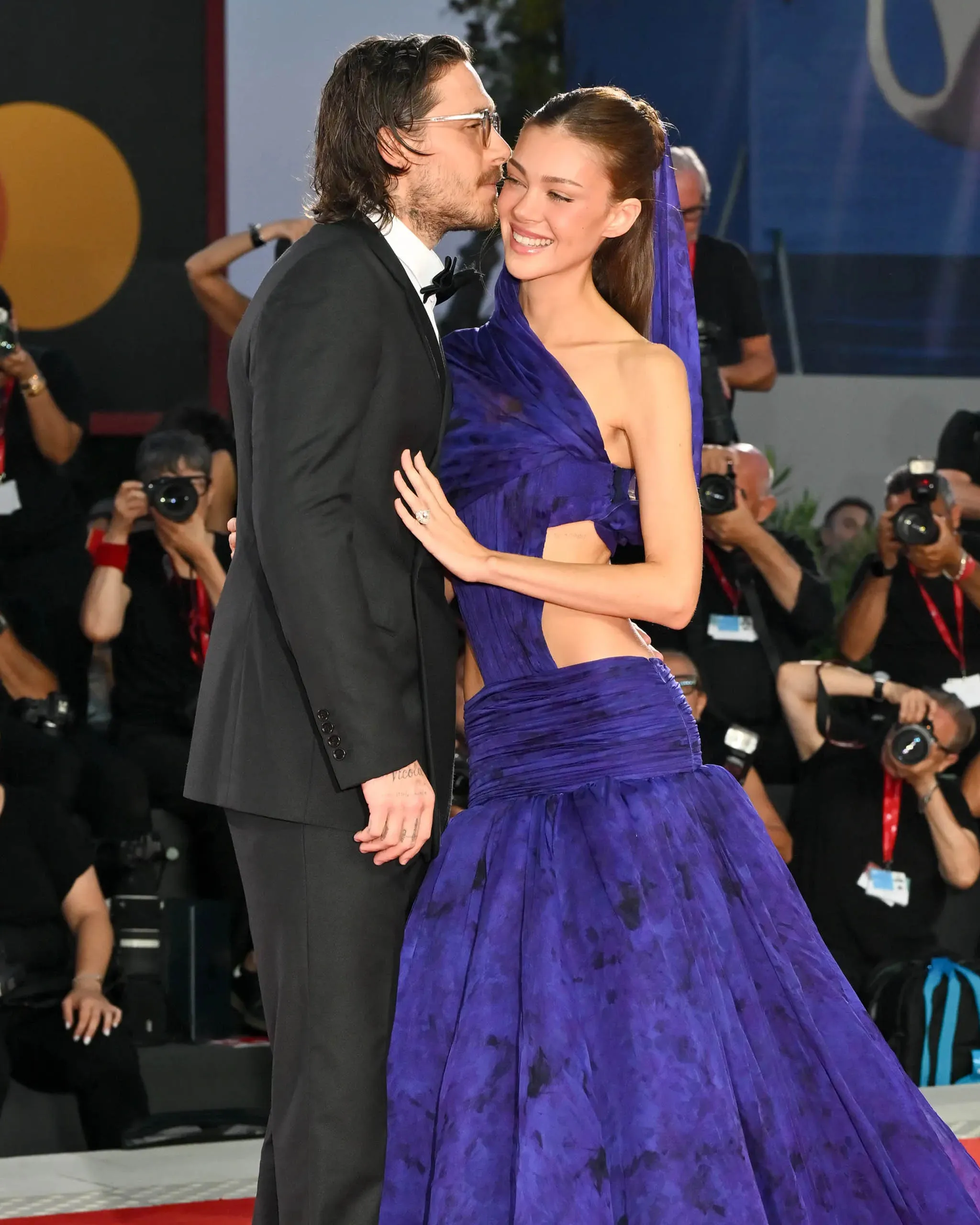 Couple on the red carpet smiling and embracing, man in a black tuxedo, woman in a purple gown amid photographers.