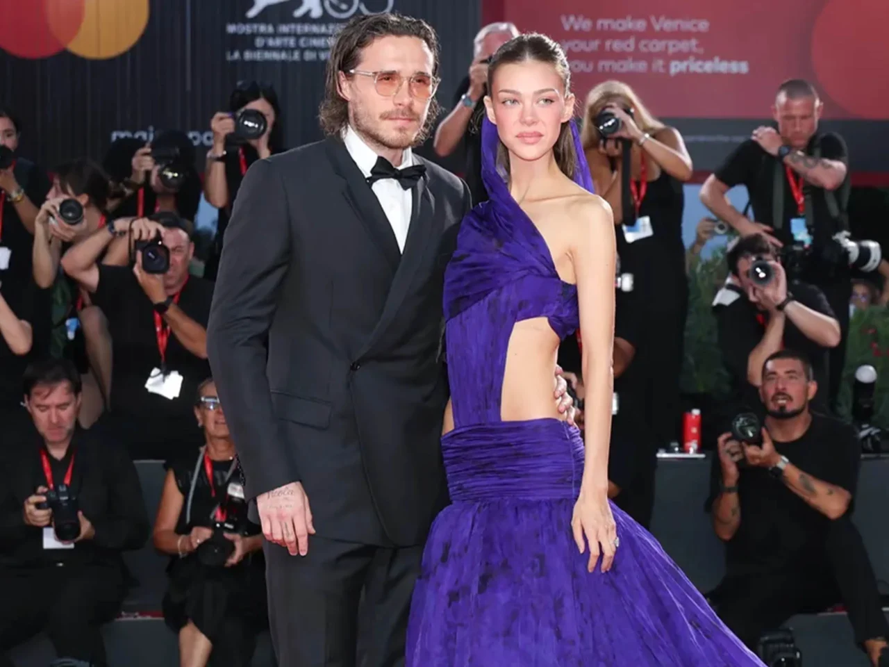 Man in a black tuxedo and a woman in a purple gown pose together on the red carpet as photographers crowd the background.