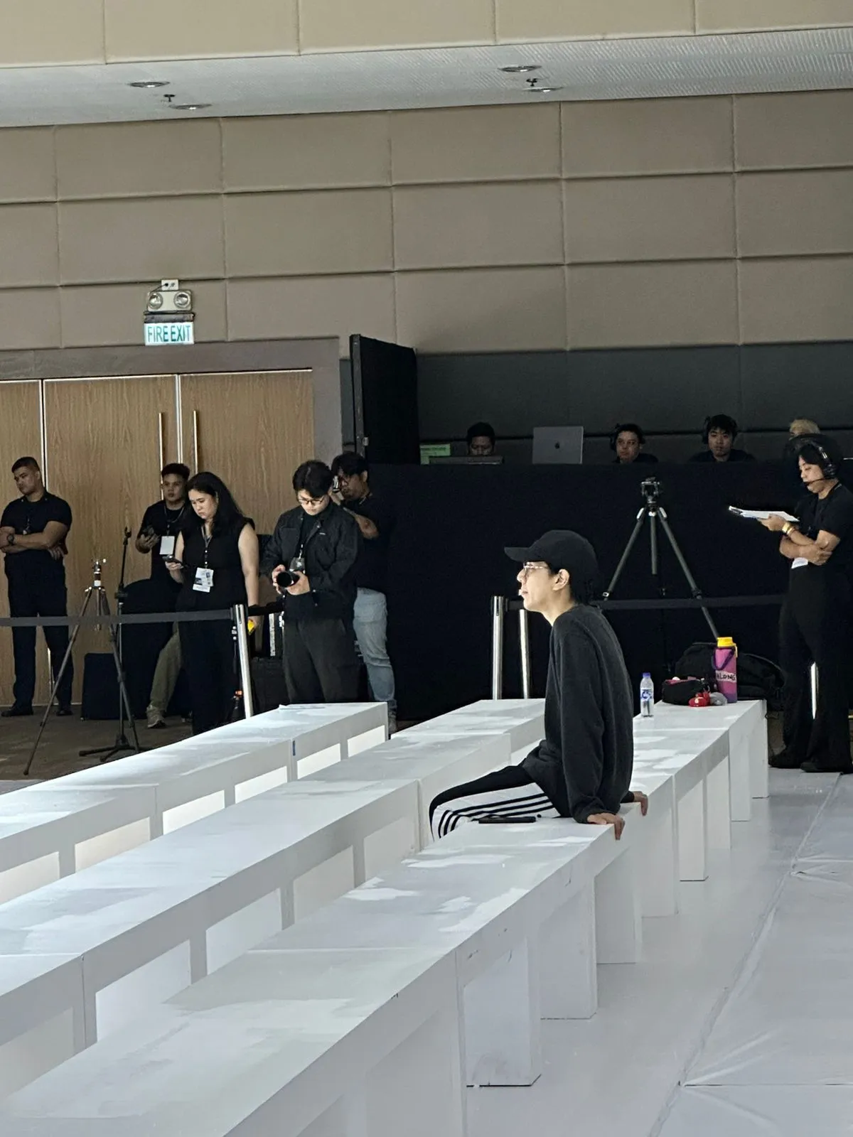 Person sits on white runway benches during a fashion show, photographers and crew behind a black barrier.