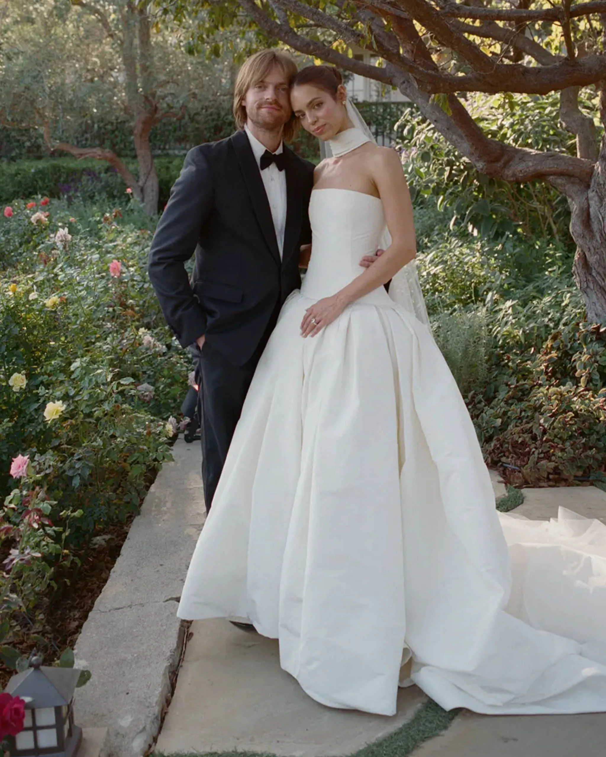 Bride in a white strapless gown with veil leaning against a groom in a black tuxedo in a garden setting.