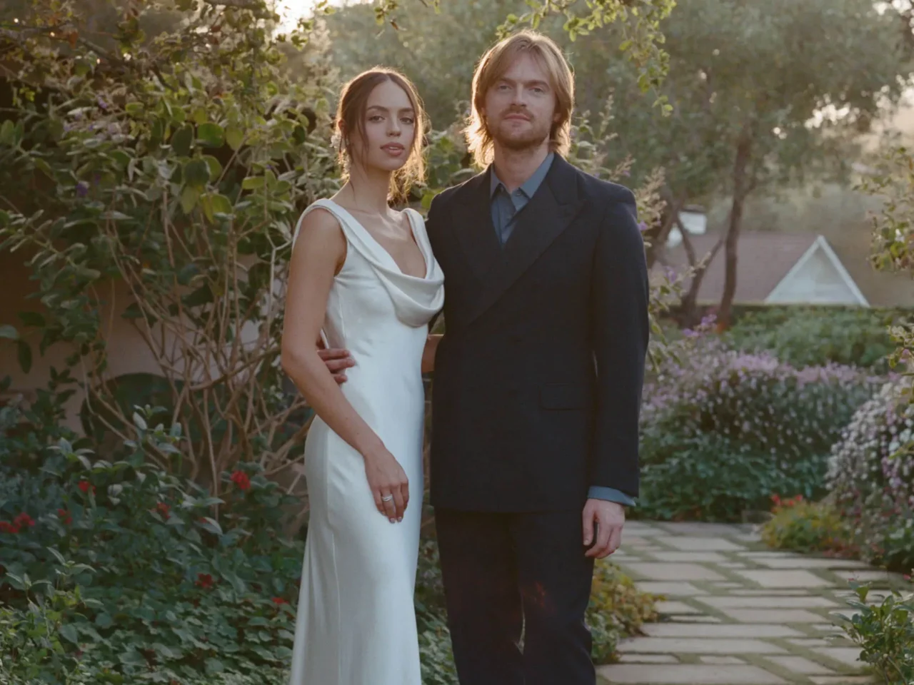 Bride and groom pose together in a garden, the bride in a white strapless gown and veil, the groom in a black tuxedo with bow tie, leaning on a tree behind colorful roses.