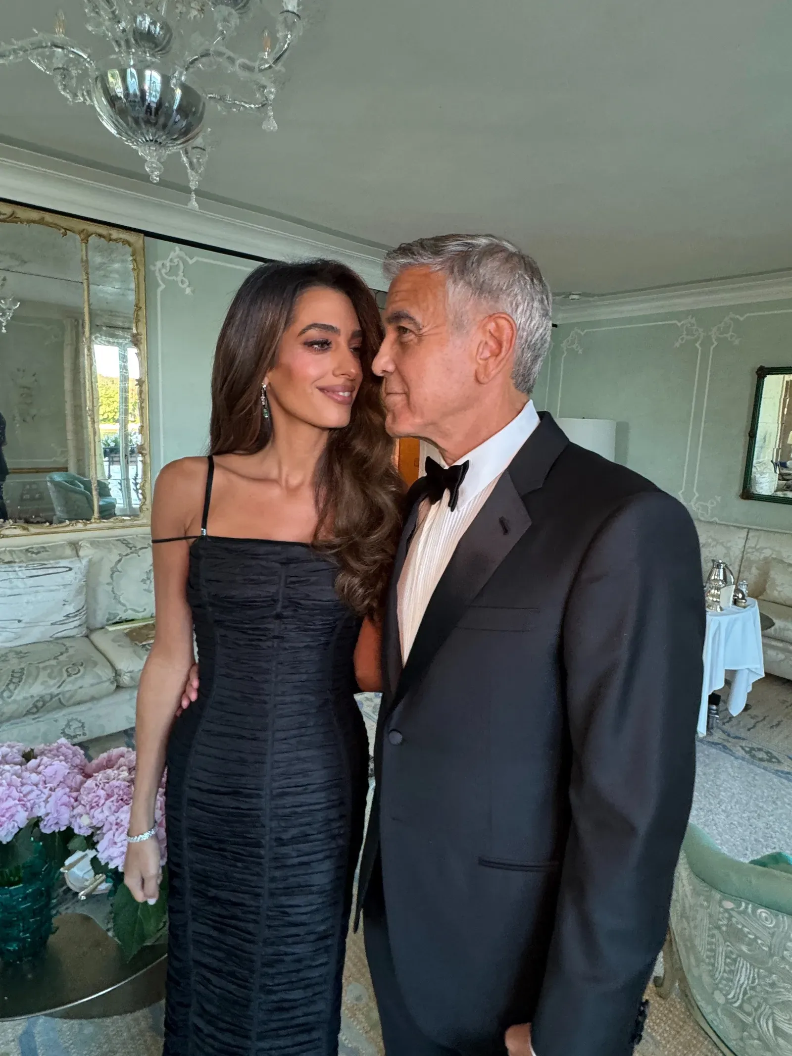 Woman in a black dress stands beside a man in a tuxedo, both smiling in an elegant room with chandeliers and mirrors in the background.