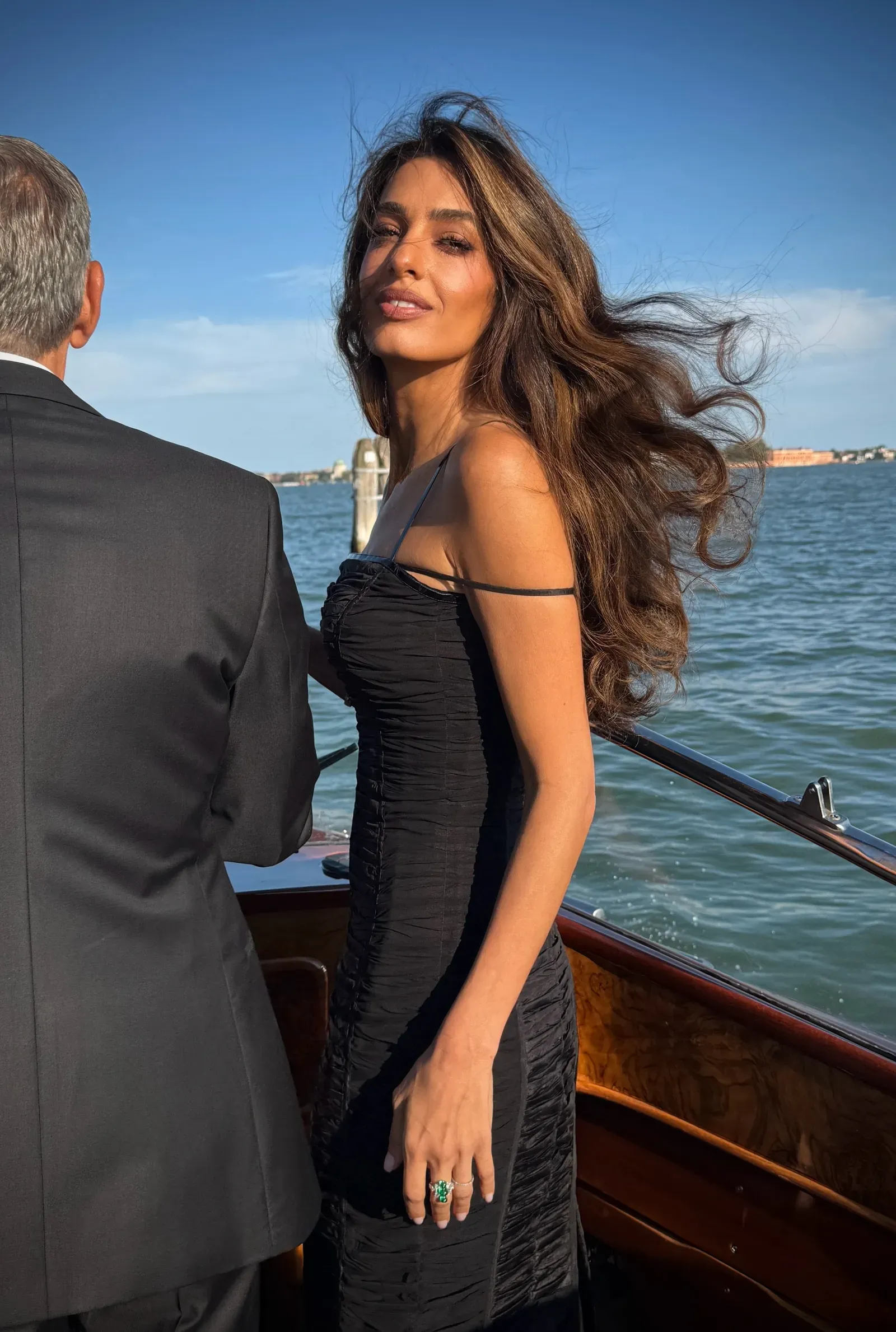 Woman in a black dress on a boat, facing the camera with wind in her hair, beside a man in a suit; water and distant shoreline in the background.