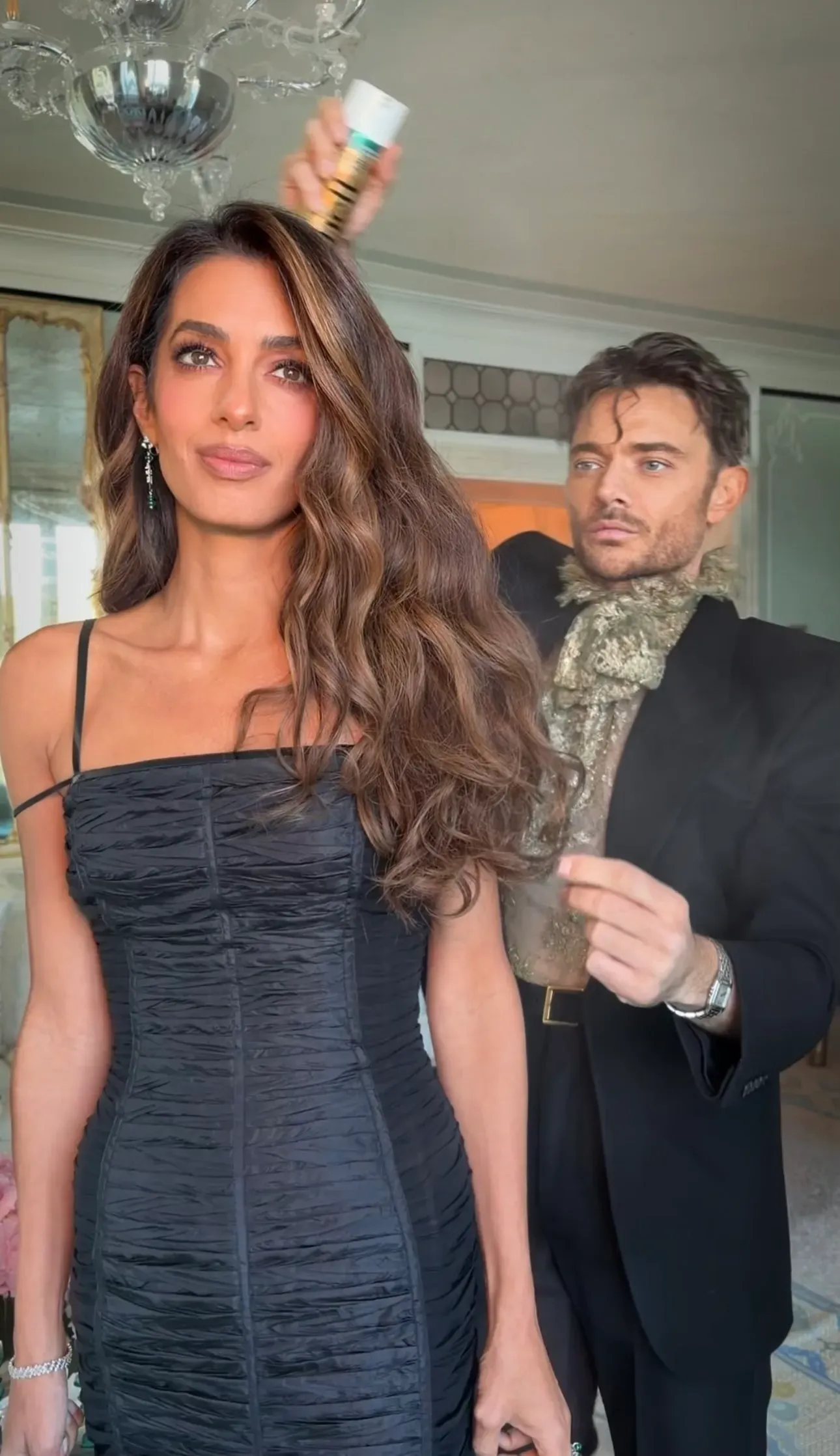 Stylist sprays hair product onto a woman with long brunette waves wearing a black dress.