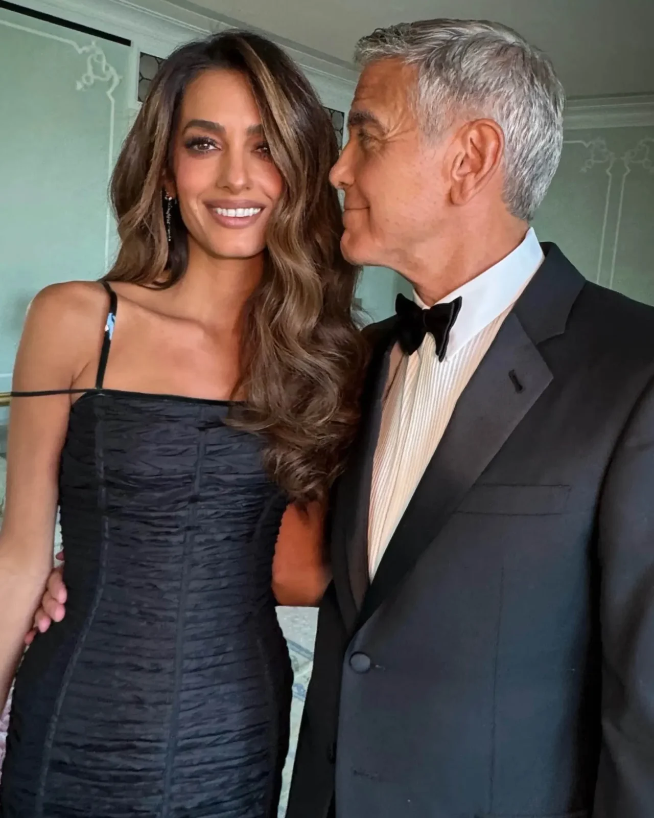 Smiling woman in a black off-shoulder dress stands next to a man in a tuxedo who is kissing her cheek in a formal setting.