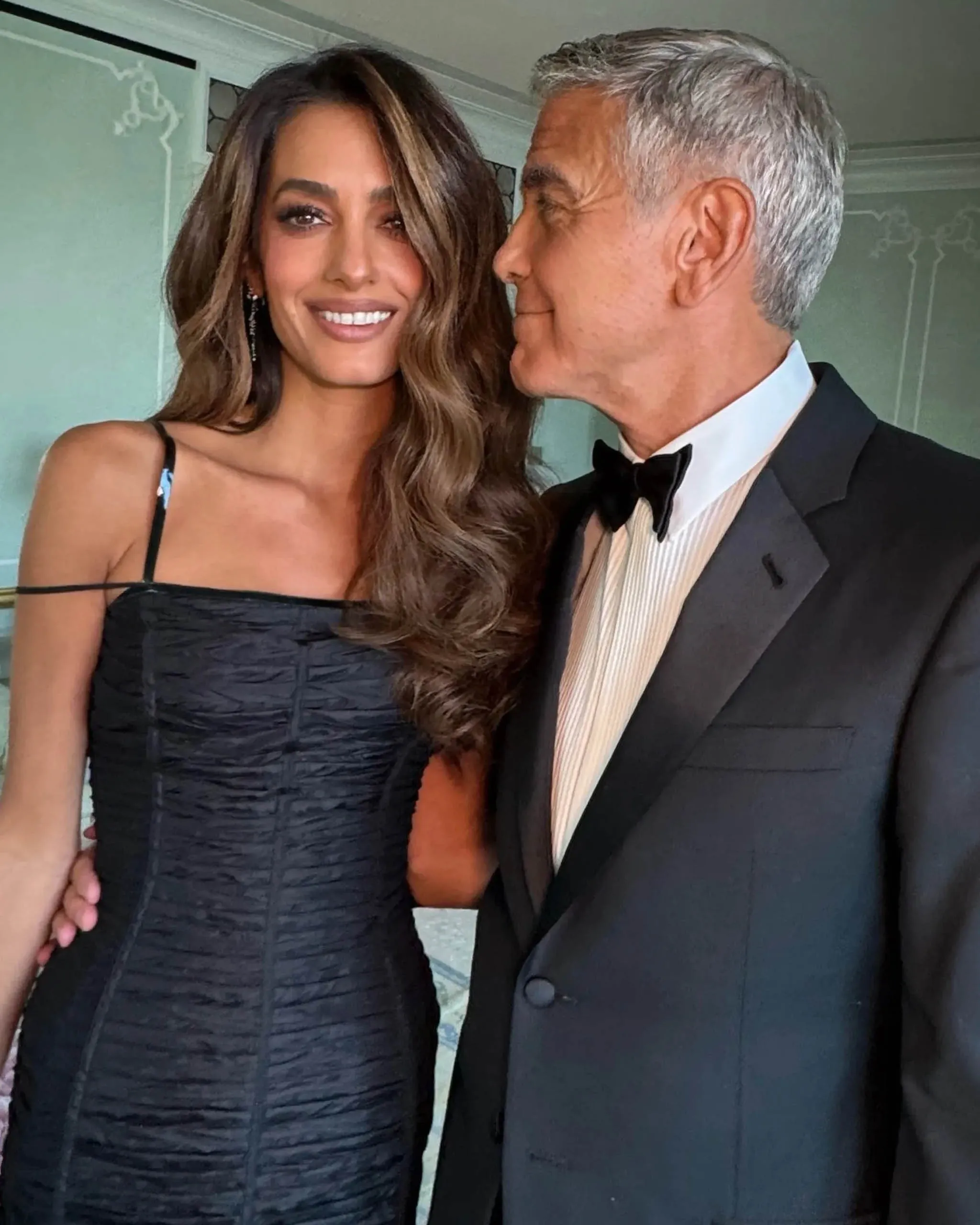 Woman in a black textured dress smiles beside a man in a tuxedo, both looking toward each other in a formal setting.