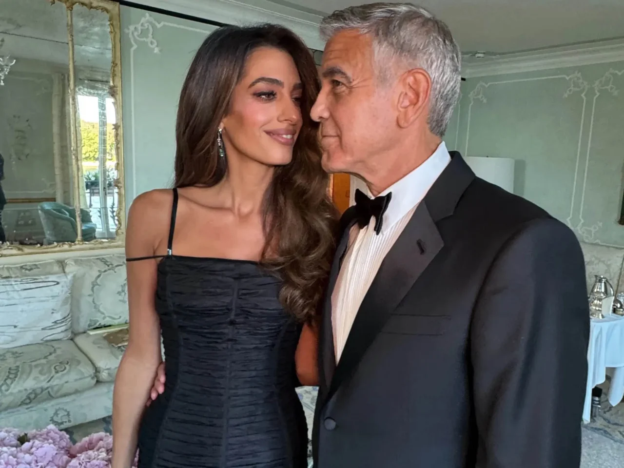 Smiling woman in a black off-shoulder dress stands next to a man in a tuxedo who is kissing her cheek in a formal setting.