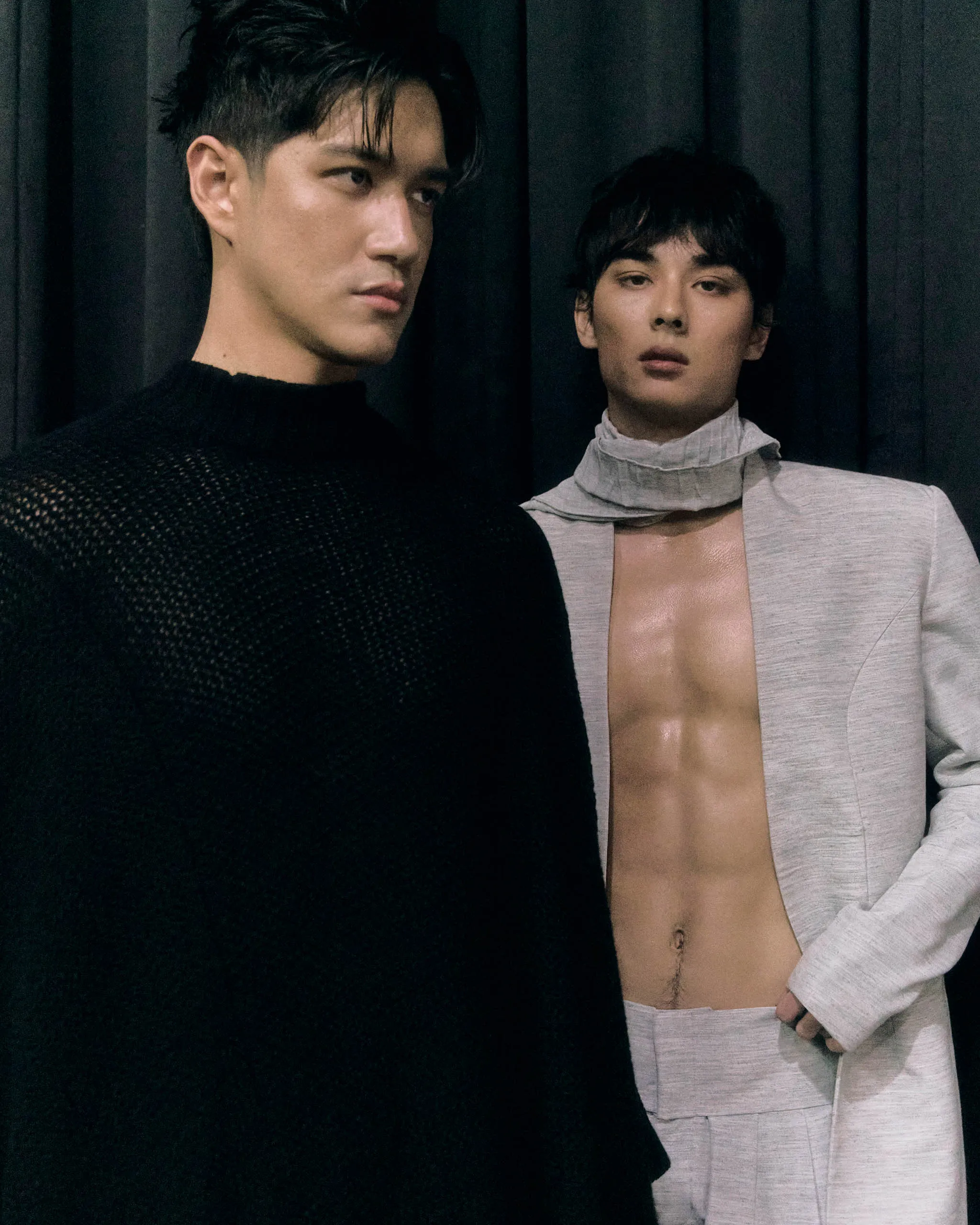 Two models stand close together against a dark curtain, one in a black knit and the other in a light gray suit with an exposed chest.