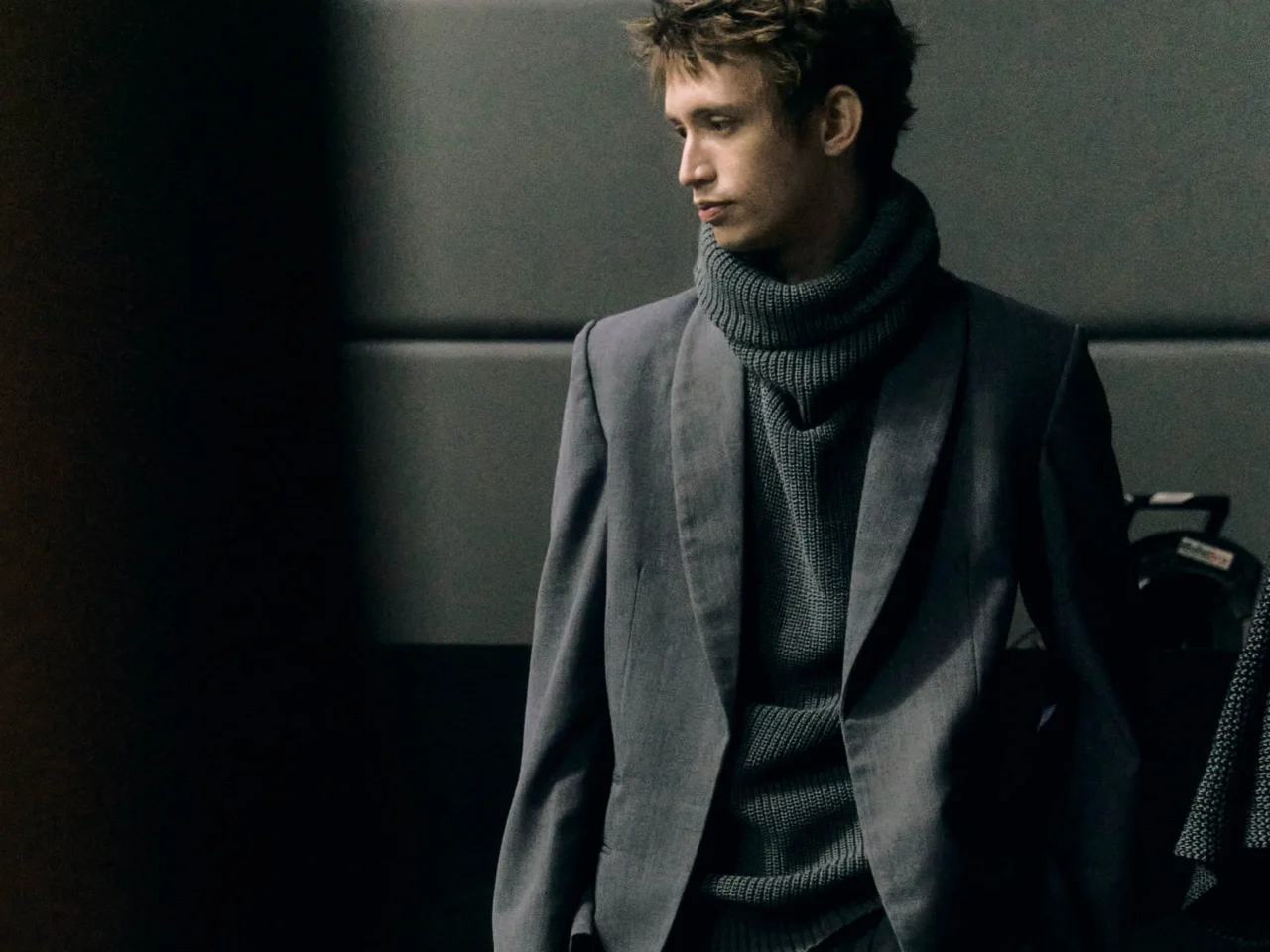 Two fashion models stand side by side wearing avant-garde outerwear: a grey textured poncho with blue fringe on the left and a dark oversized coat on the right, against a plain backdrop.