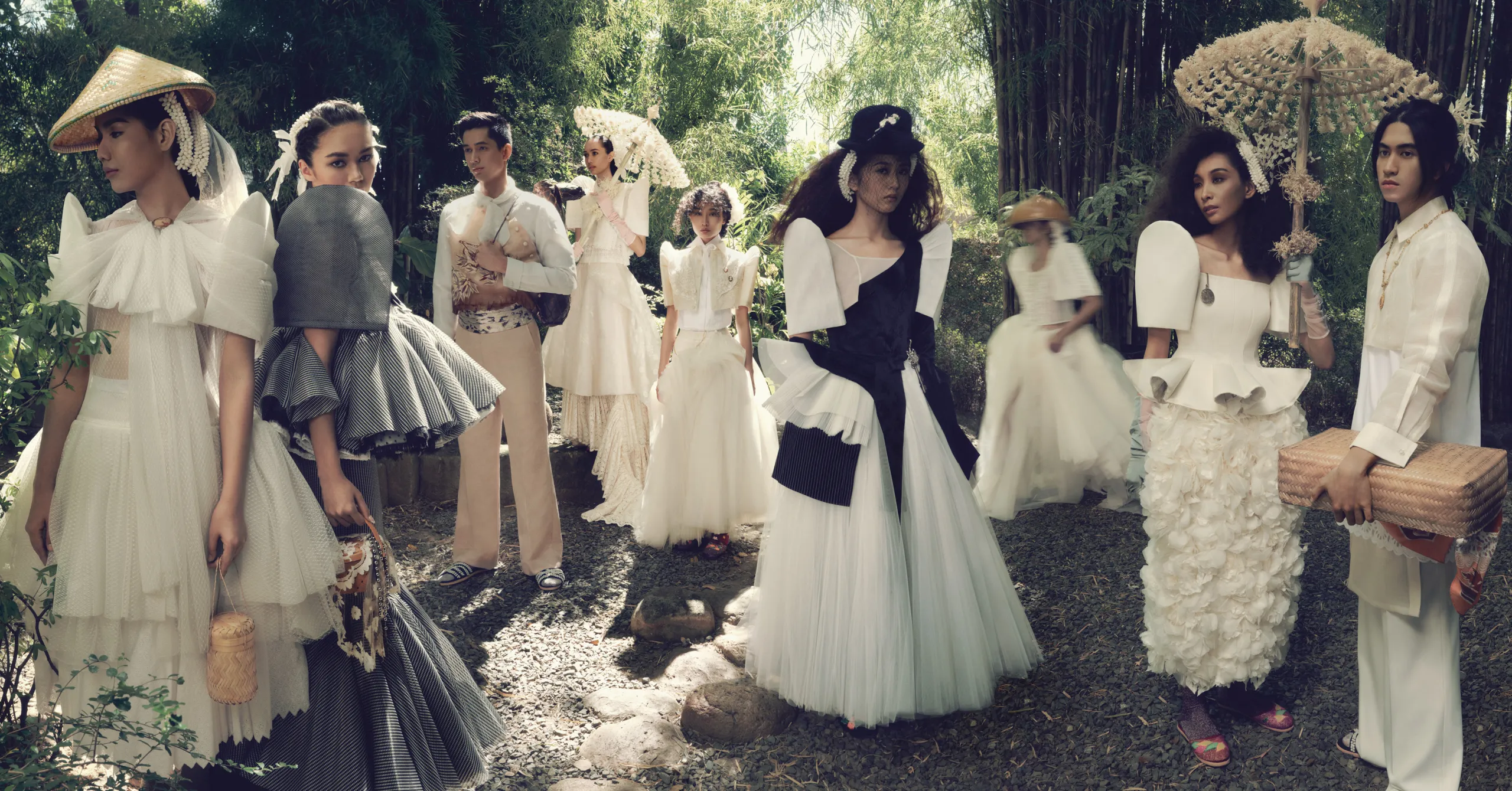 Group of models in white couture gowns posing in a sunlit garden with oversized hats and parasols.