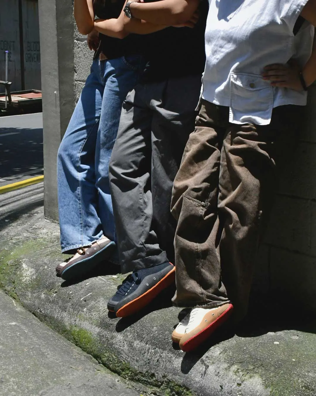 Four people standing side by side against a concrete wall on a city sidewalk, showing their lower bodies and colorful sneakers.