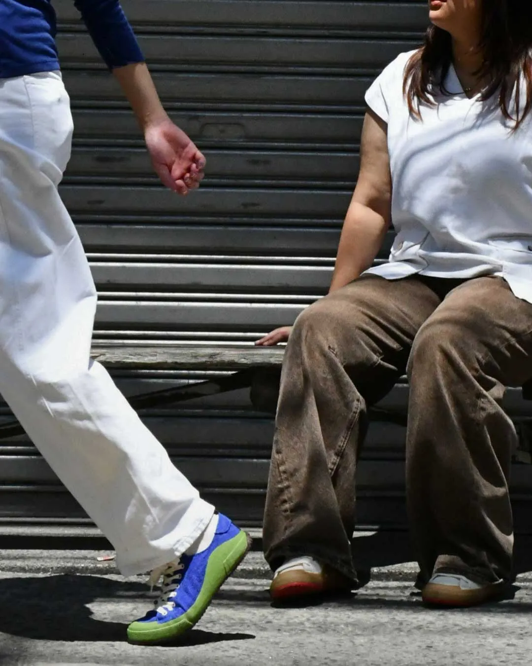 Person in a white shirt and brown pants sits on a metal bench as another person in white pants and bright blue‑green sneakers walks by.