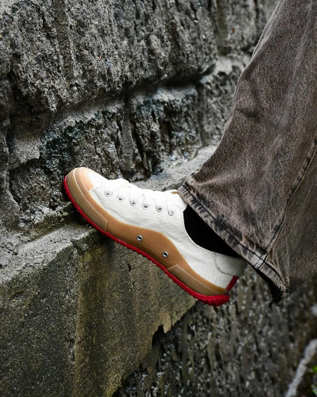White canvas sneaker with a red outsole pressed against a rough stone wall, pant leg visible in brown jeans.