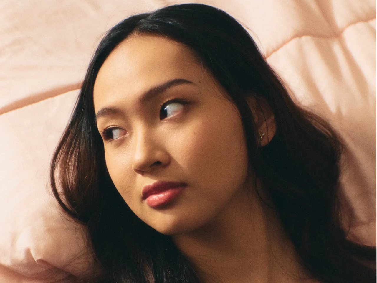 Close-up of a young woman with dark hair looking to her left against a soft peach background, neutral expression.