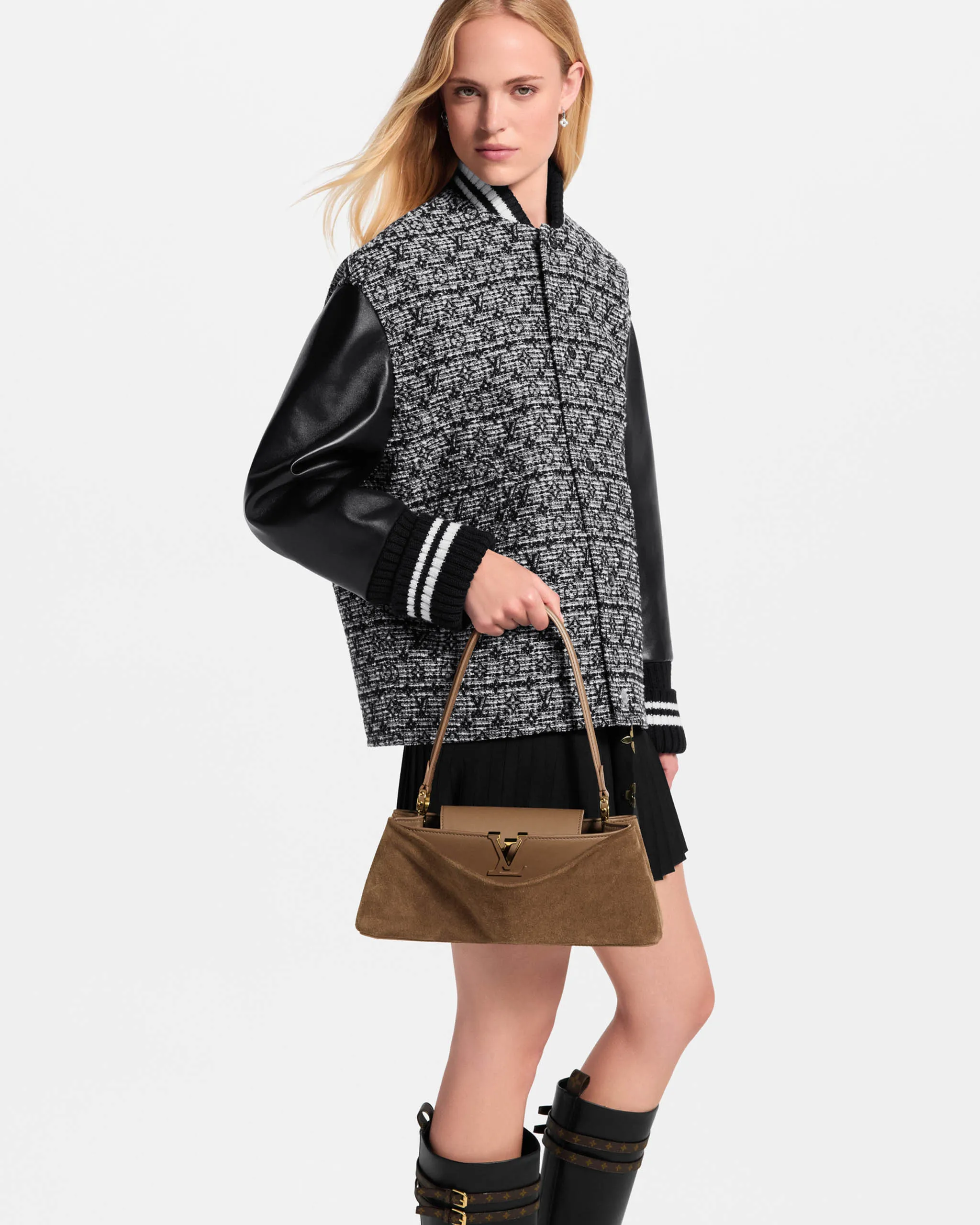 Model in a gray textured jacket with black leather sleeves, holding a tan handbag against a white backdrop.