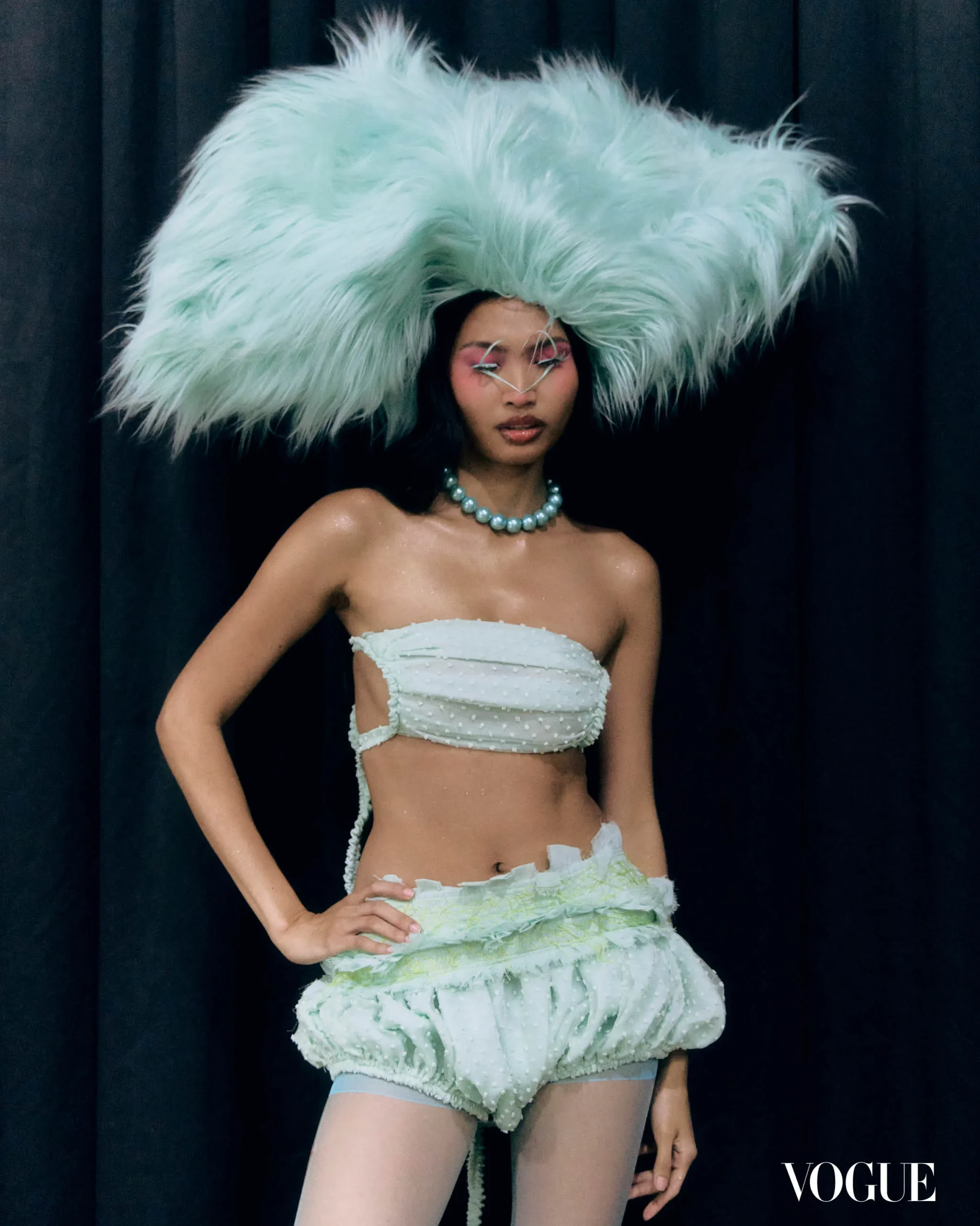 Fashion model wearing an oversized pale blue feathered headdress, pearl necklace, and light strapless top against a dark backdrop (Vogue).