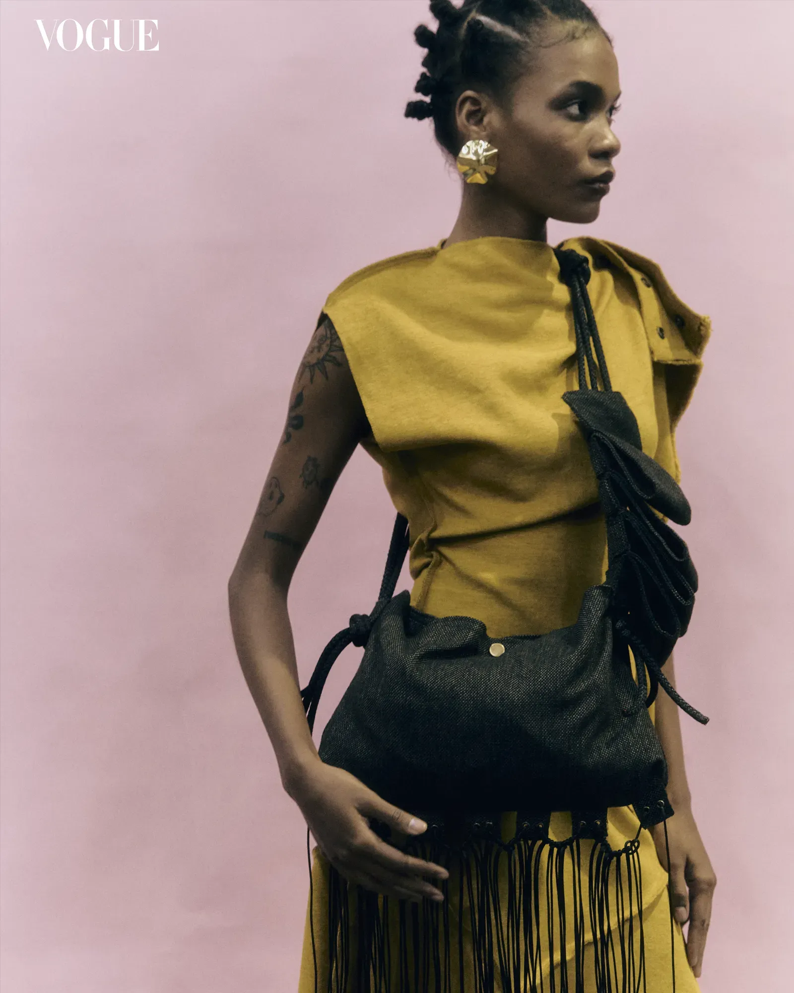 Model in a mustard yellow sleeveless dress with a black fringe bag, gold geometric earring, and braided hair against a pink backdrop; Vogue logo visible in the top left corner.
