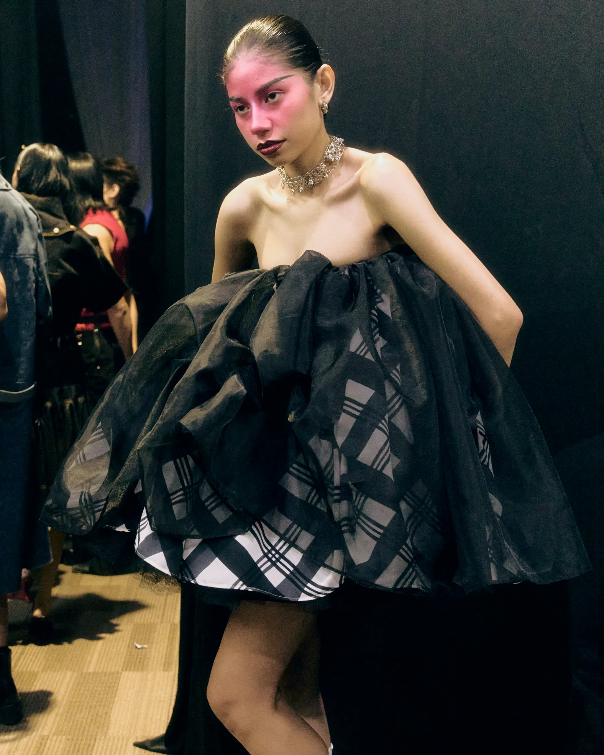 Backstage fashion model in a dramatic black voluminous gown with off-shoulder silhouette and a chunky silver necklace, pink-hued makeup, and dark lipstick.
