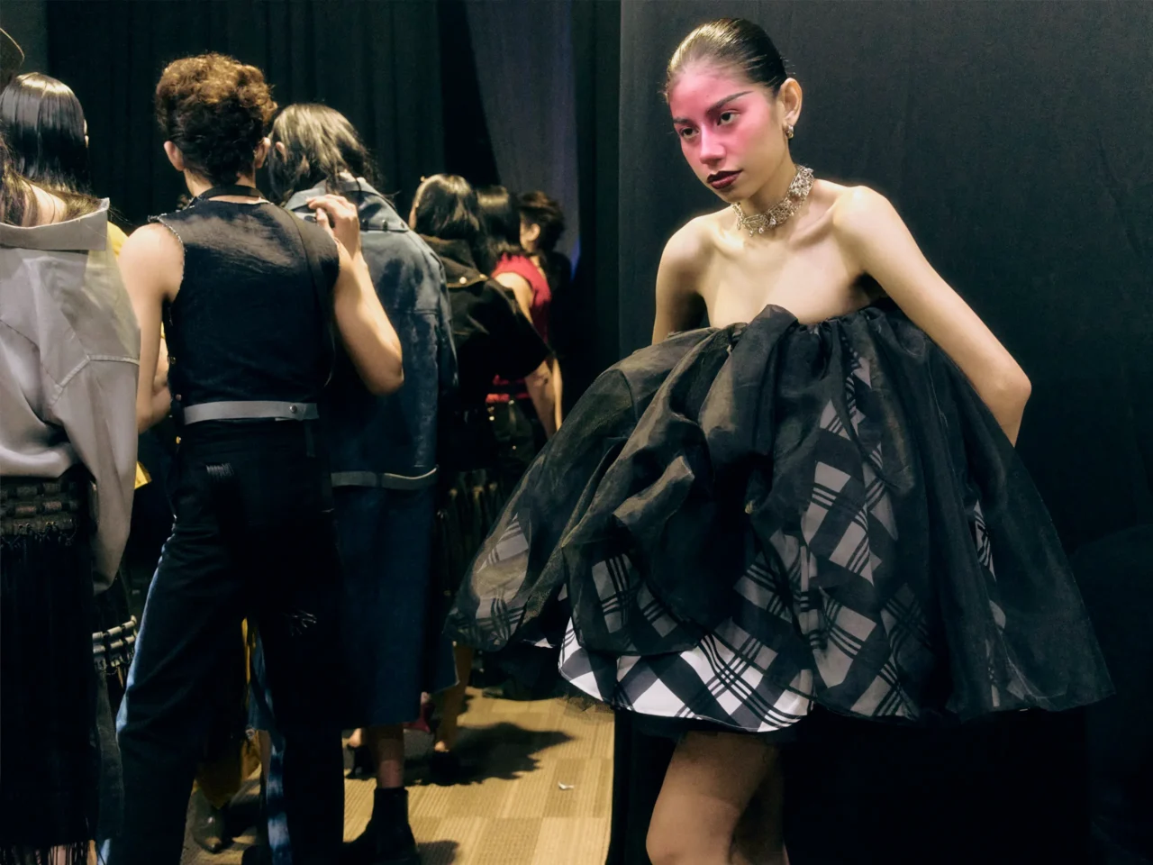 Backstage fashion show scene with a model in a dark, voluminous gown on the right and crew members prepping in the background.