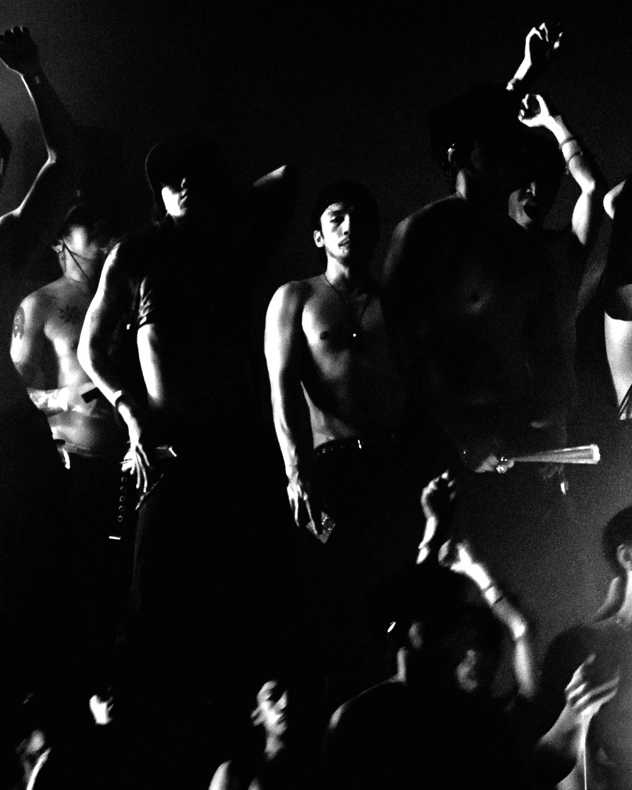 Crowd of shirtless men dancing in a dark club, with raised arms and high-contrast lighting.