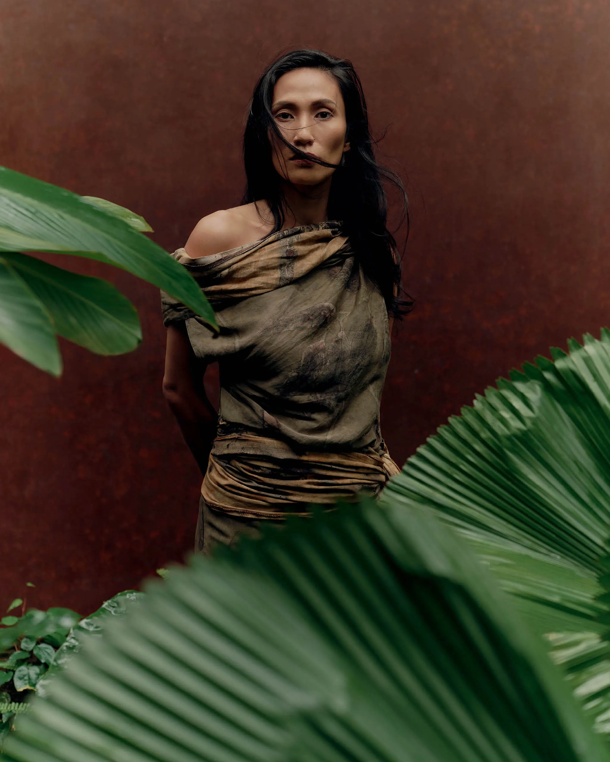 Woman standing outdoors in a tan and brown wrap against a rust-colored wall, with large green tropical leaves partially in the foreground.