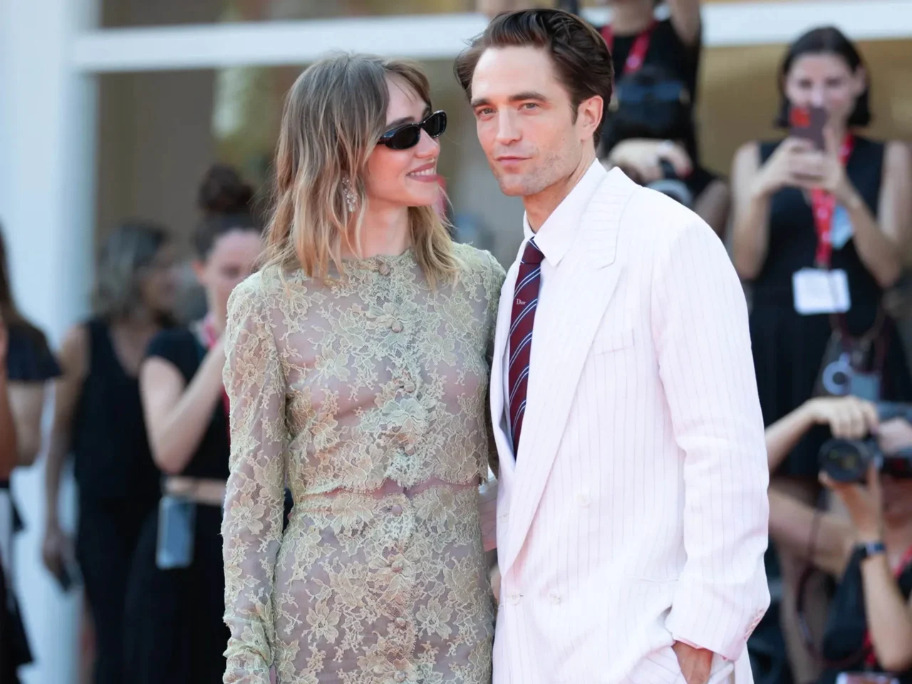Couple posing at a formal event; woman in pale green lace dress with sunglasses, man in a light pink pinstripe suit and burgundy tie.