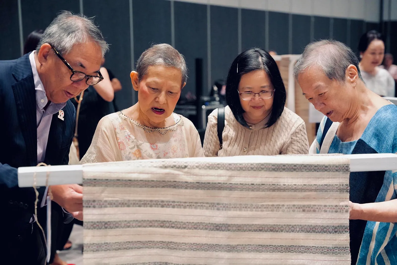 Four older adults closely examine a striped woven textile on a loom in a workshop setting, discussing its pattern.