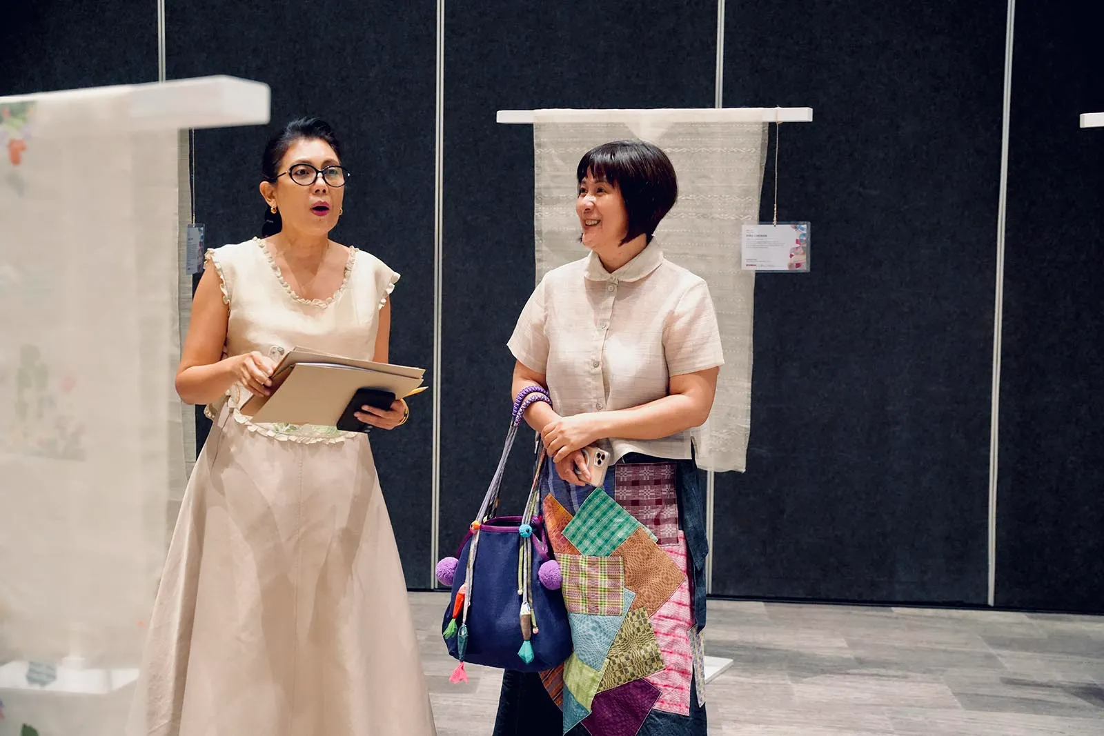 Two women at an exhibition talk: one in a cream dress with glasses holding folders, the other smiling with a colorful patchwork bag next to her.