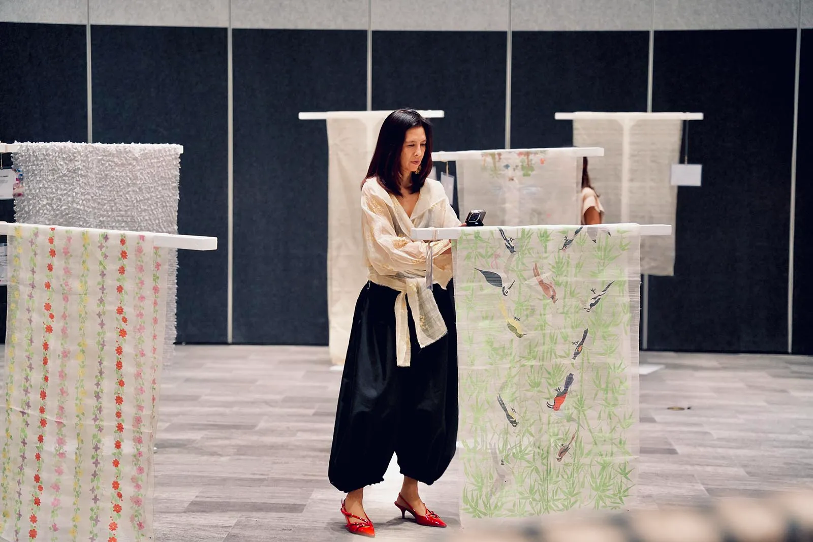 Woman in a cream blouse and black wide-leg pants with red shoes stands at a display of hanging fabrics in a showroom/galleriespace.