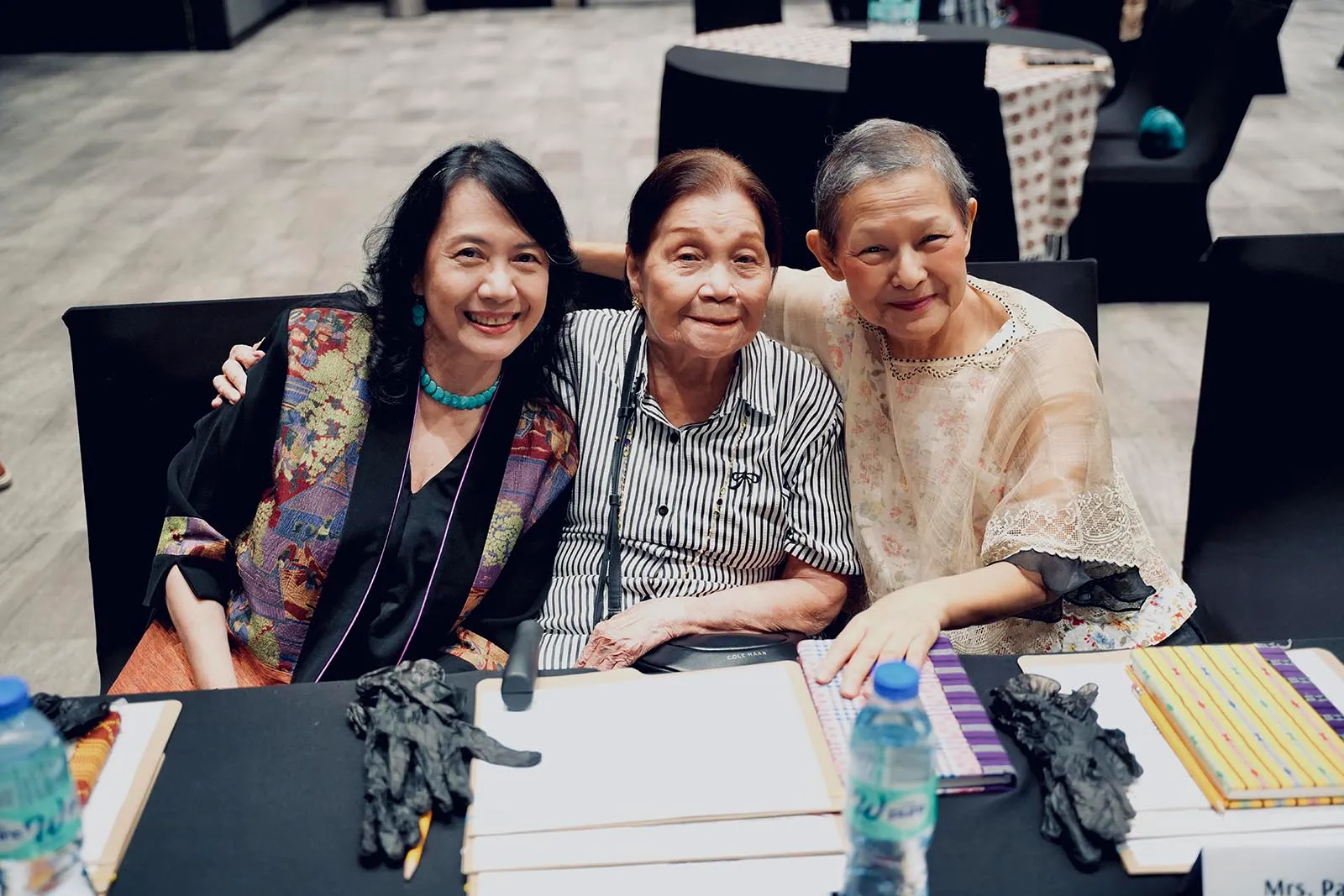 Three women sit closely together at a table, smiling for the camera at an indoor event.