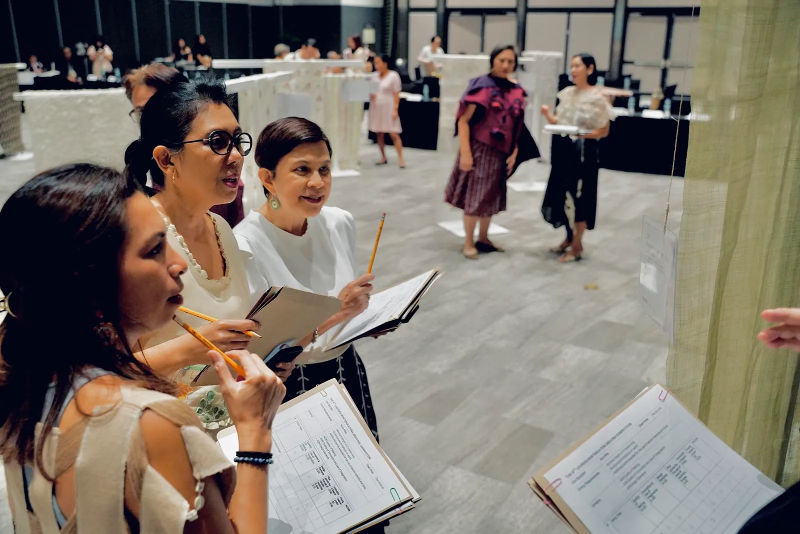 Three women with notebooks and pencils review documents at an indoor exhibition or workshop.