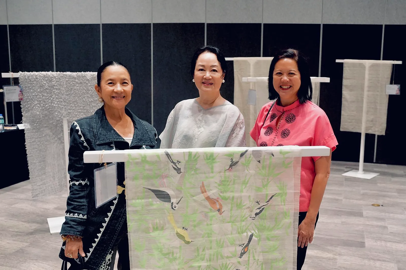Three women pose in a gallery holding a light textile with birds among green plants.
