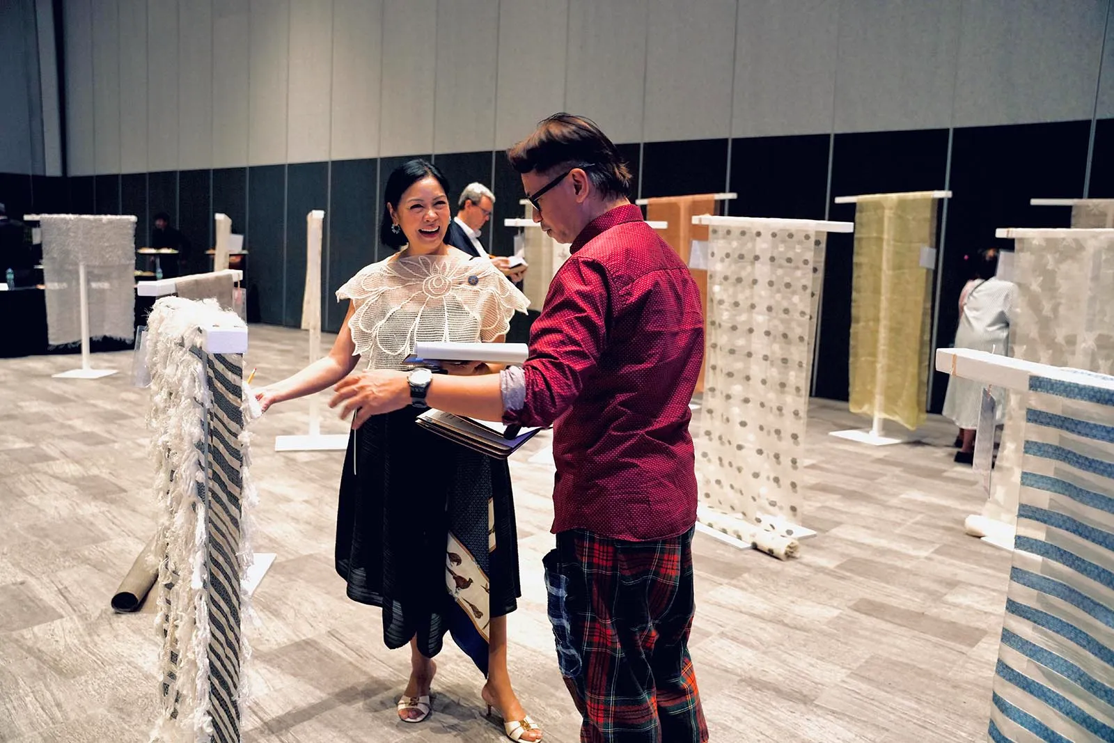 Two people discuss fabric samples at a textile exhibition, with racks of fabrics around them.