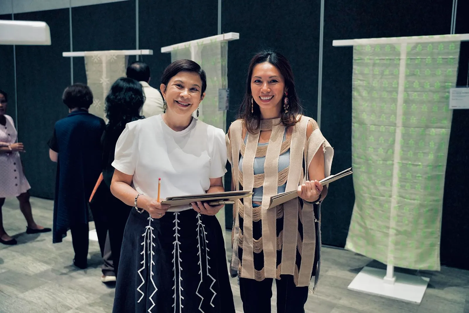 Two women smiling and holding folders at a textile exhibition, with fabric samples on display behind them.