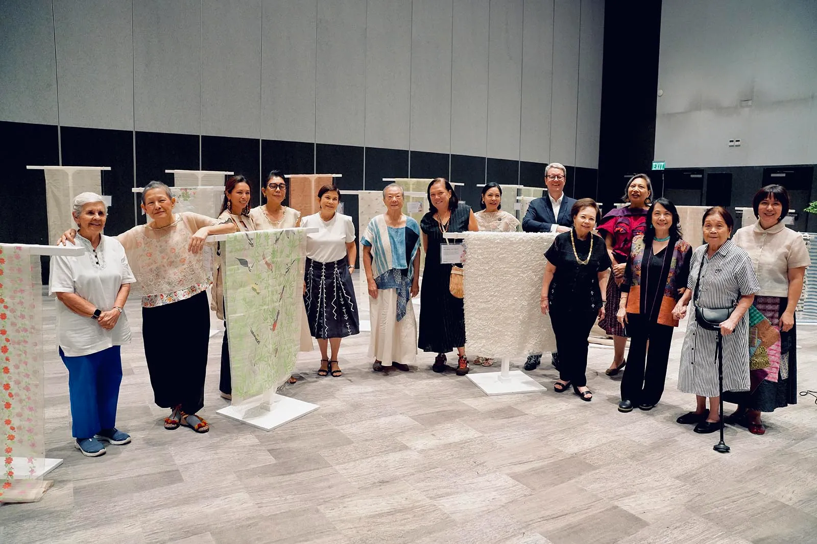 Group of people standing in a textile exhibition, posing behind fabric swatches on display stands in a large hall, smiling at the camera.