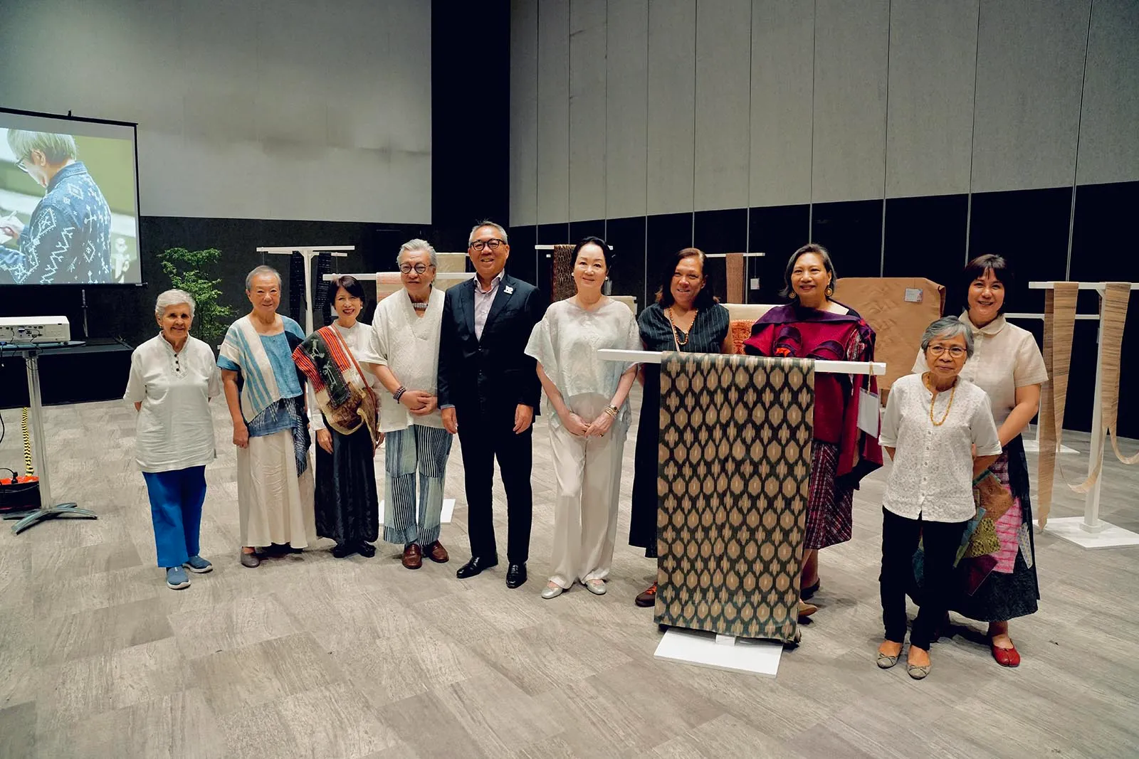 Group of diverse adults posing for a photo in a textile showroom, with woven fabric samples and a projector screen in the background.