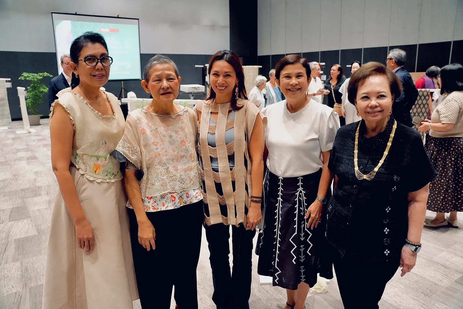 Five women posing together at a formal indoor event, wearing varied light to dark outfits; a projection screen is in the background.