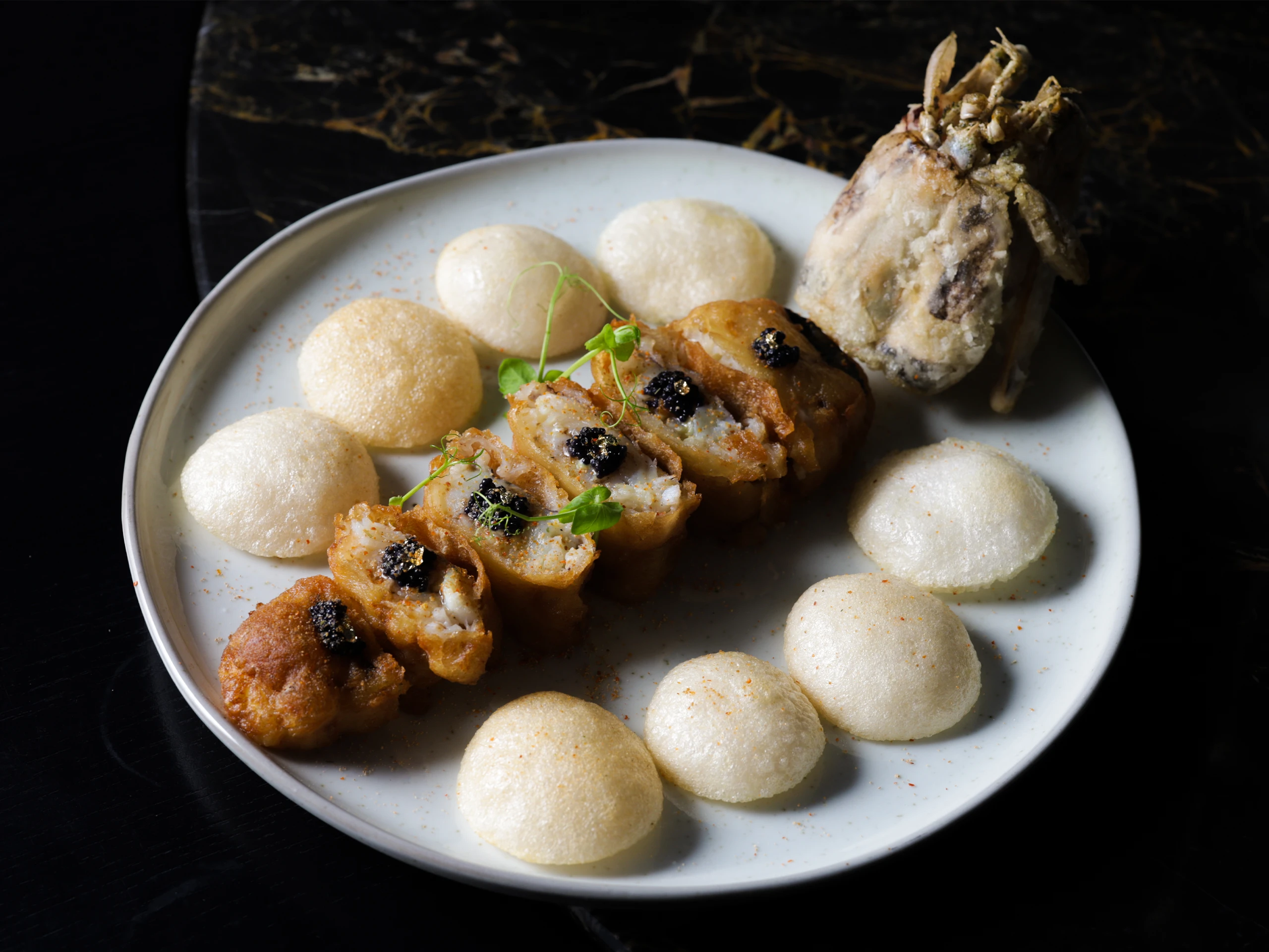 Plate of fried shrimp with crispy rounds and a fried fish tail, garnished with microgreens on a dark surface.