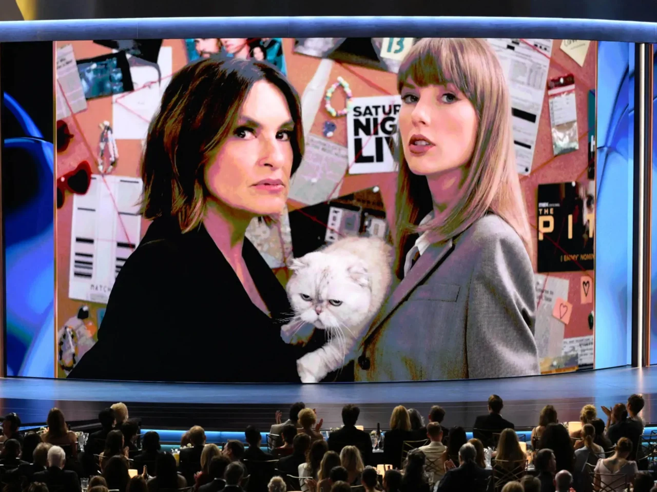 Two women stand close together holding a white cat, in front of a bulletin-board backdrop, shown on a large screen in a theater.