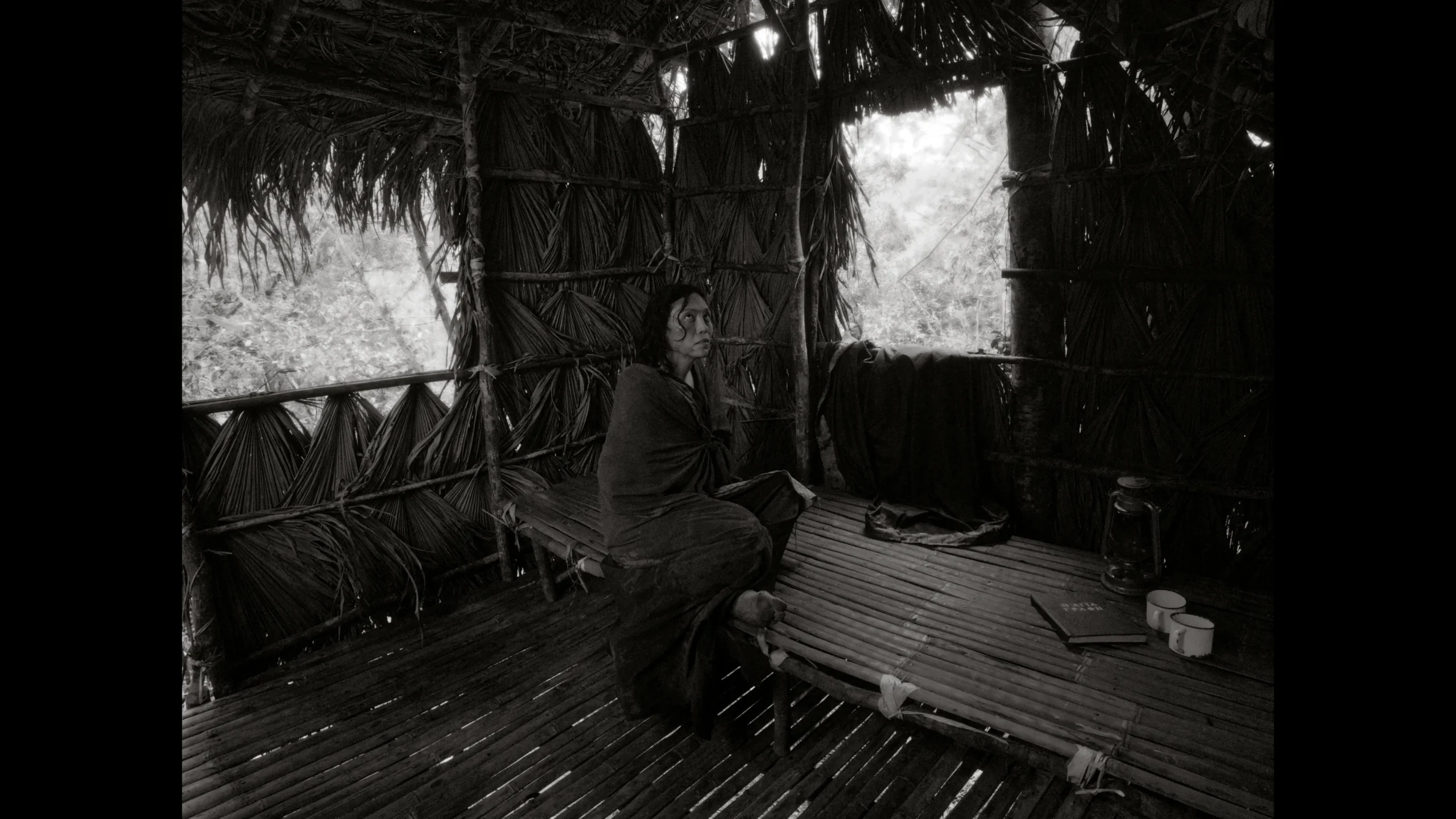Woman seated inside a thatched bamboo hut, wrapped in a cloak, looking to the left.